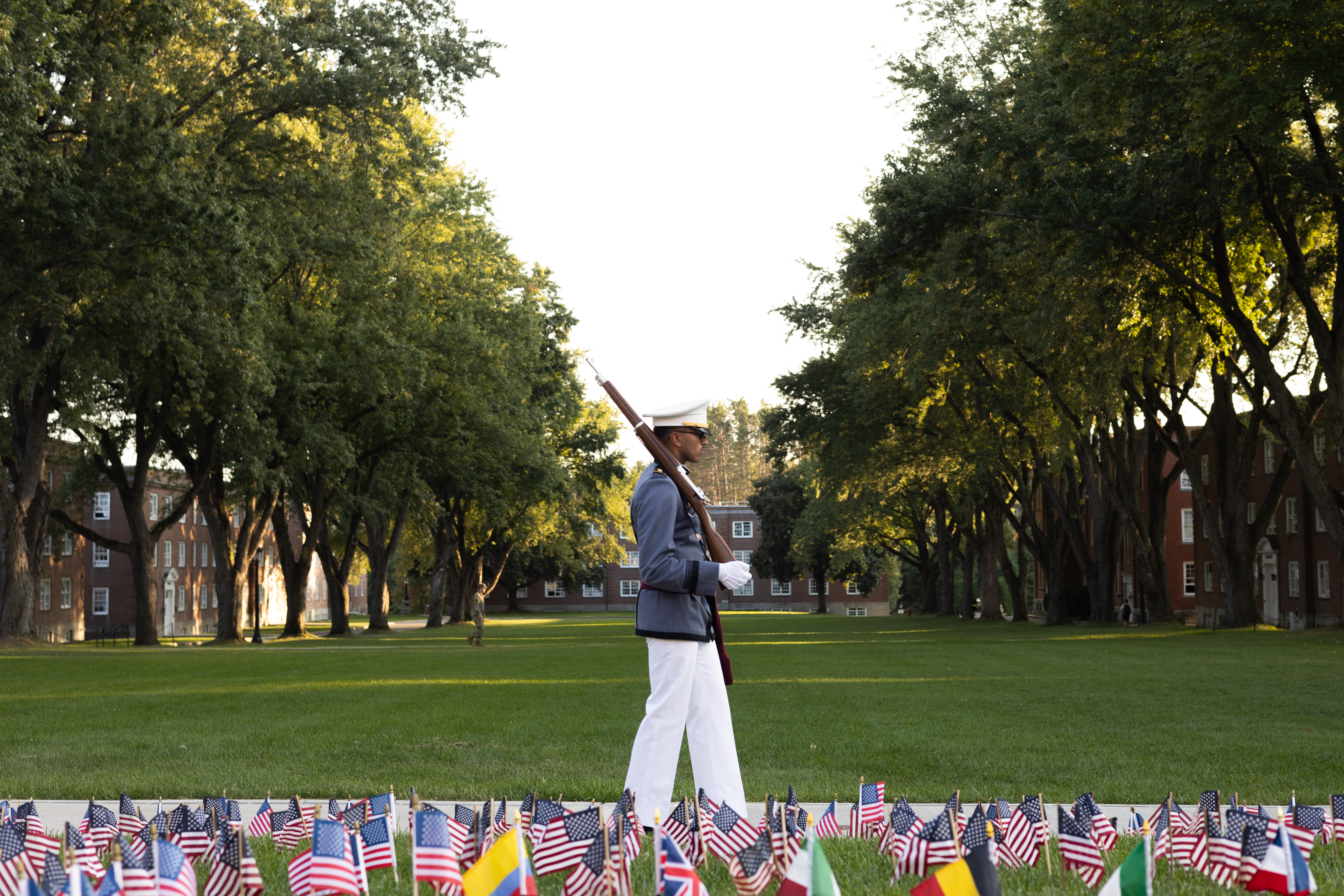 Flags are planted in the ground on the Upper Parade Ground immediately in the foreground; just behind them, a cadet walks along the Tour Strip with a rifle over his right shoulder, and a view of the Upper Parade Ground behind him.