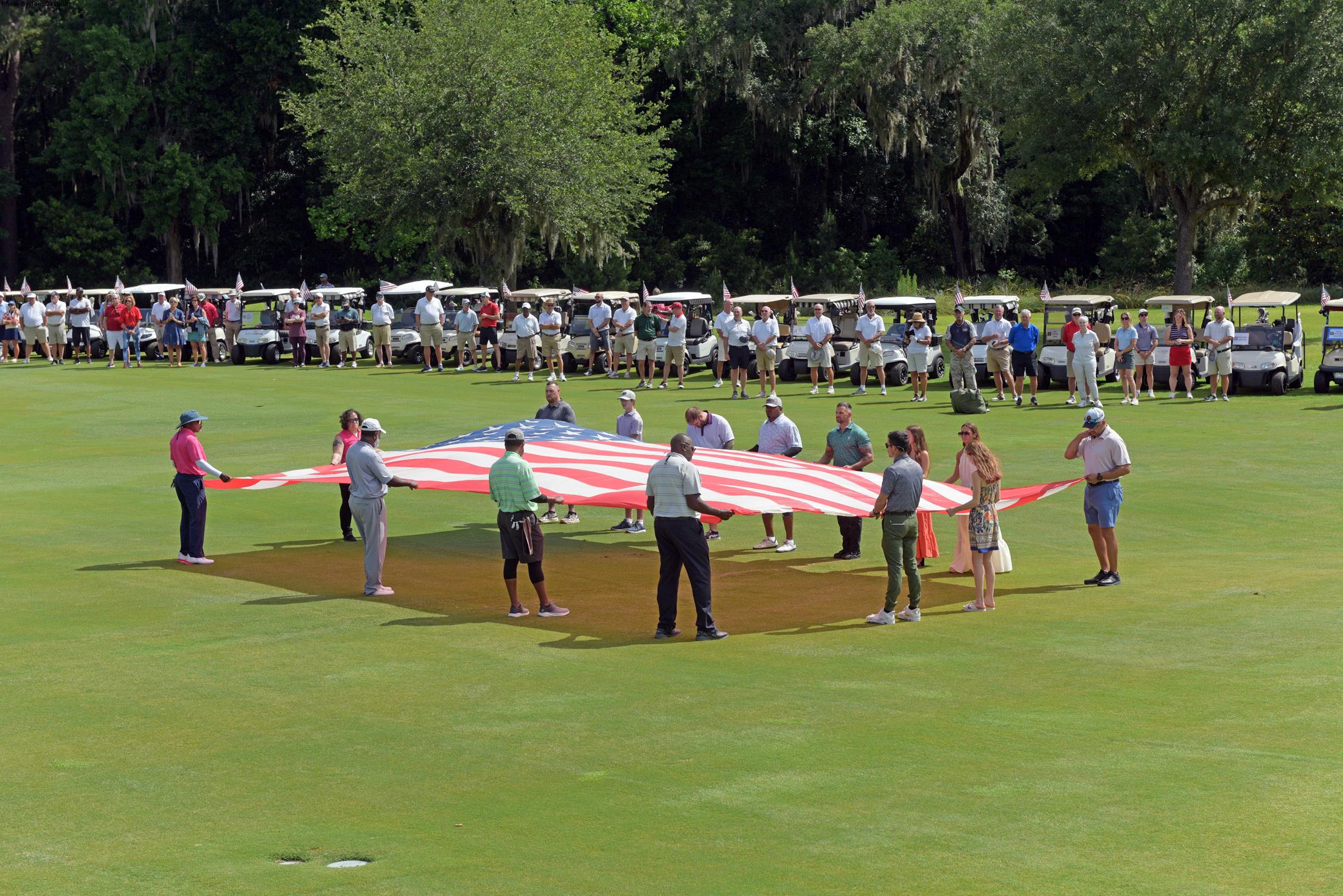 A group of people hold the American flag over a fairway green, while a line of golf carts observes from behind.