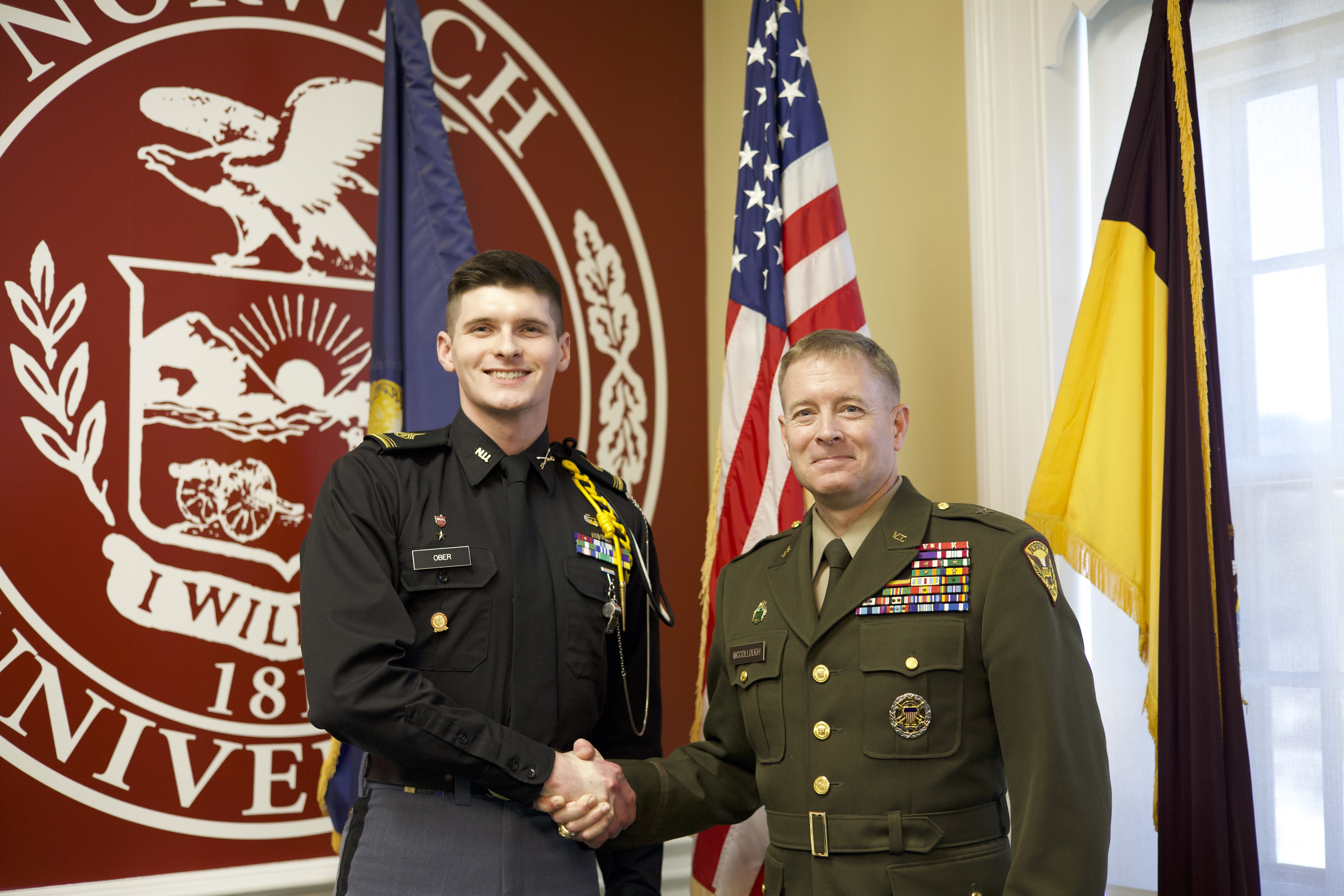 BG William F. McCollough, VSM, Commandant and Vice President for Student Affairs, Norwich Class of 1991 and C/1SG Matthew S. Ober '26 shaking hands in front of the Norwich University logo
