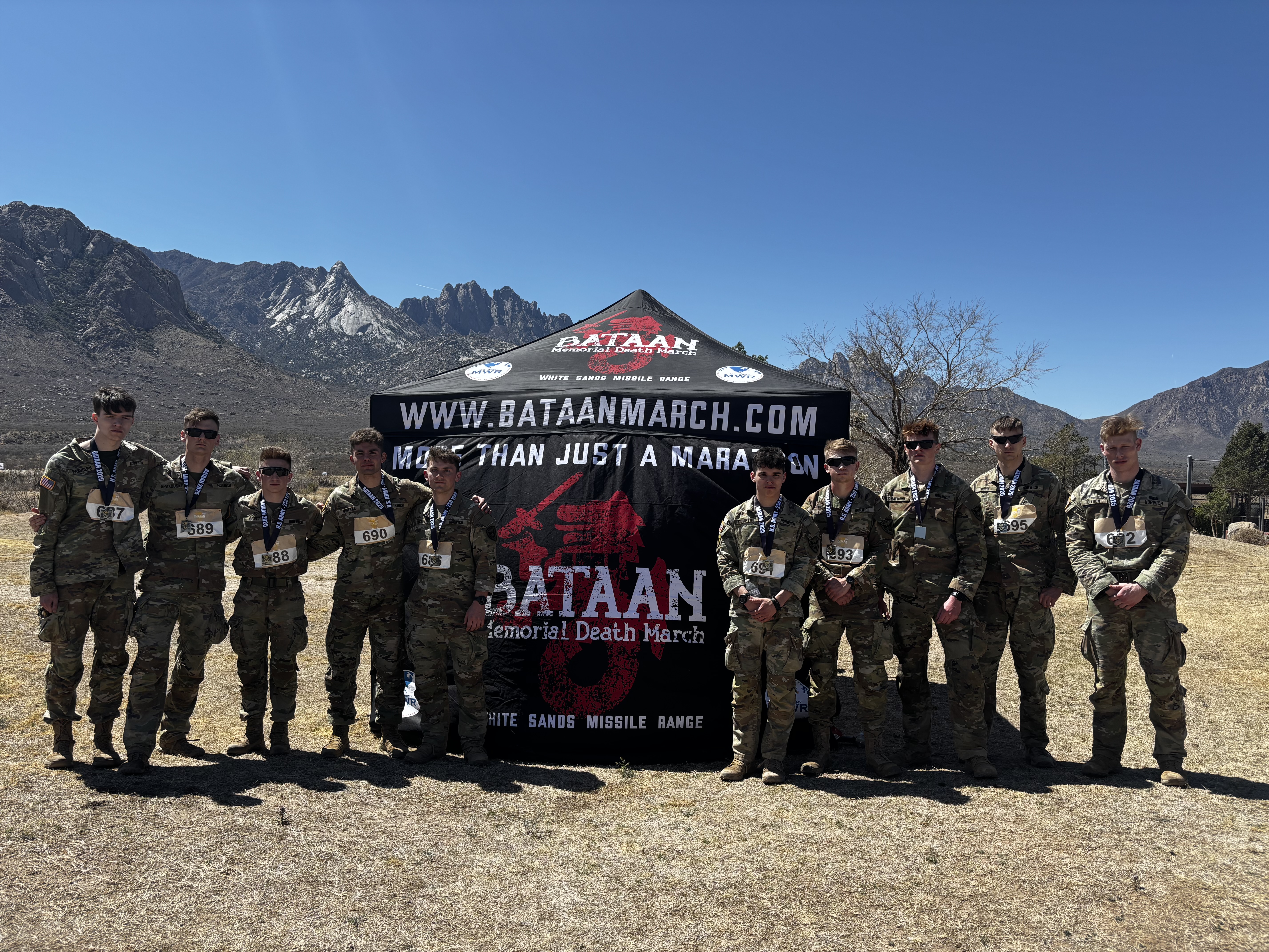The Norwich University cadets who participated in the event stand shoulder-to-shoulder in front of a branded tent in the desert with a mountain range in the background.