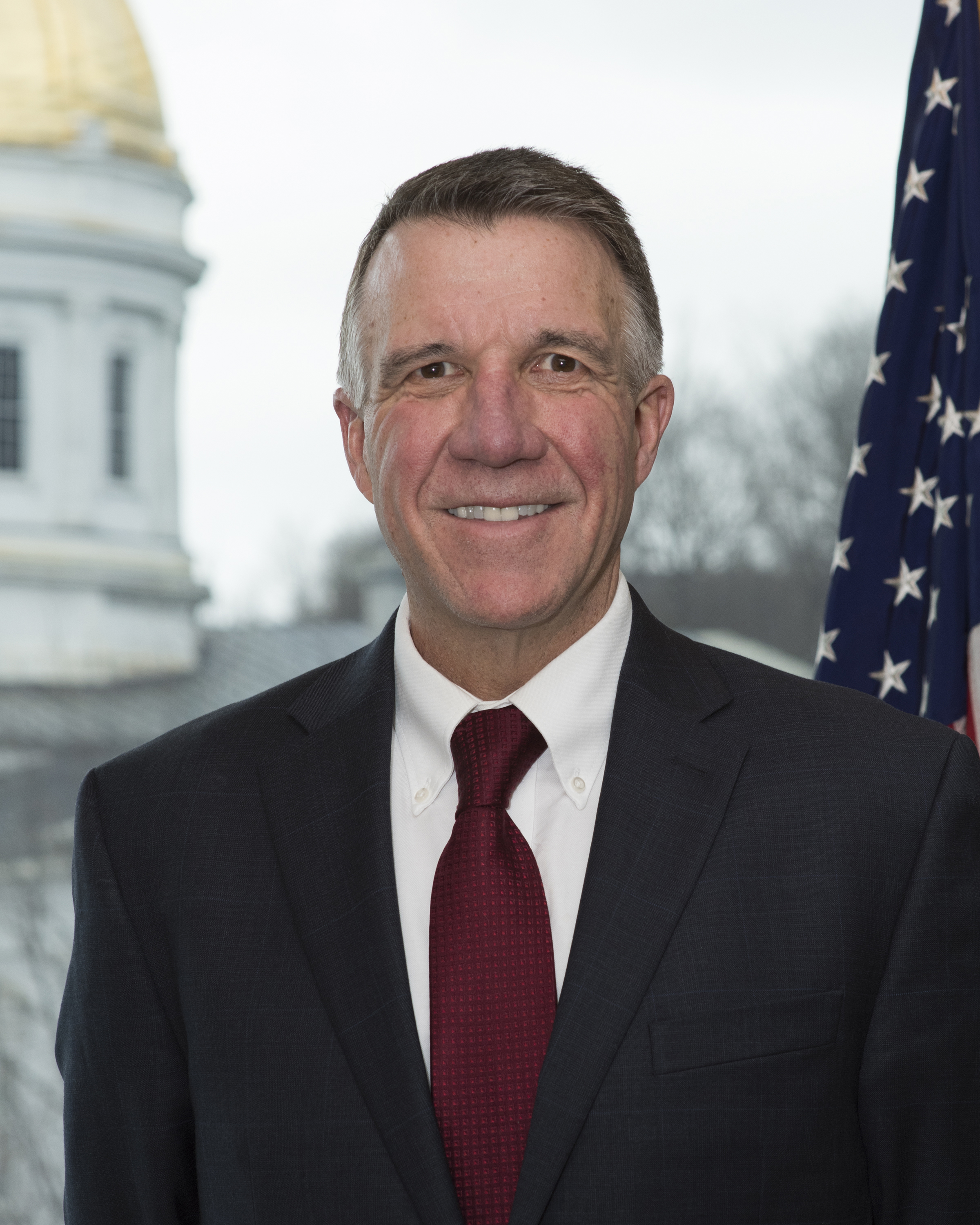A formal headshot of Governor Phil Scott in a suit flanked by an American flag behind his left shoulder, and the capitol building in the distance behind his right.