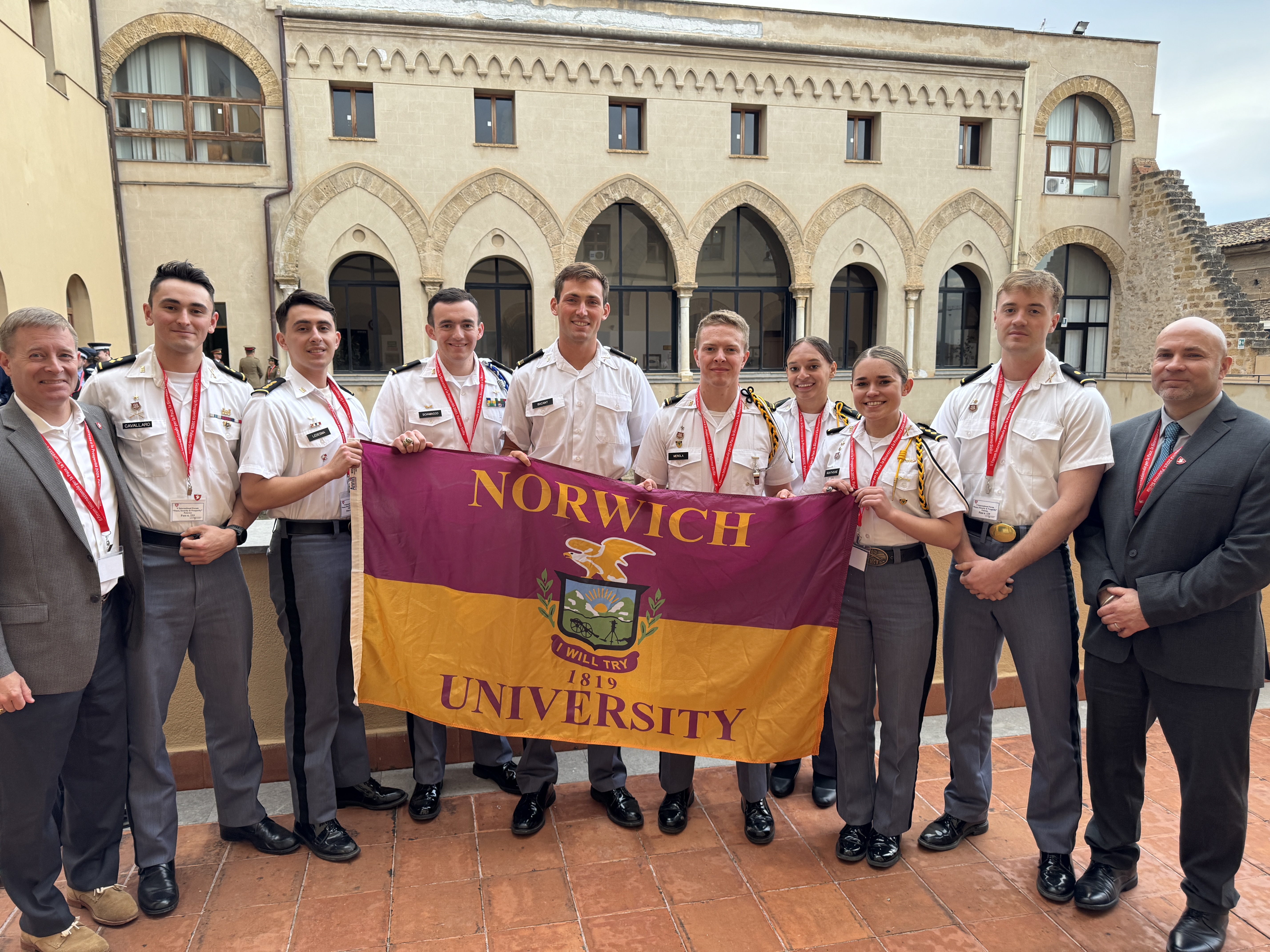 Norwich cadets stand shoulder-to-shoulder holding a Norwich flag, flanked on each end by Norwich faculty. They are standing in a courtyard with a building in the background.