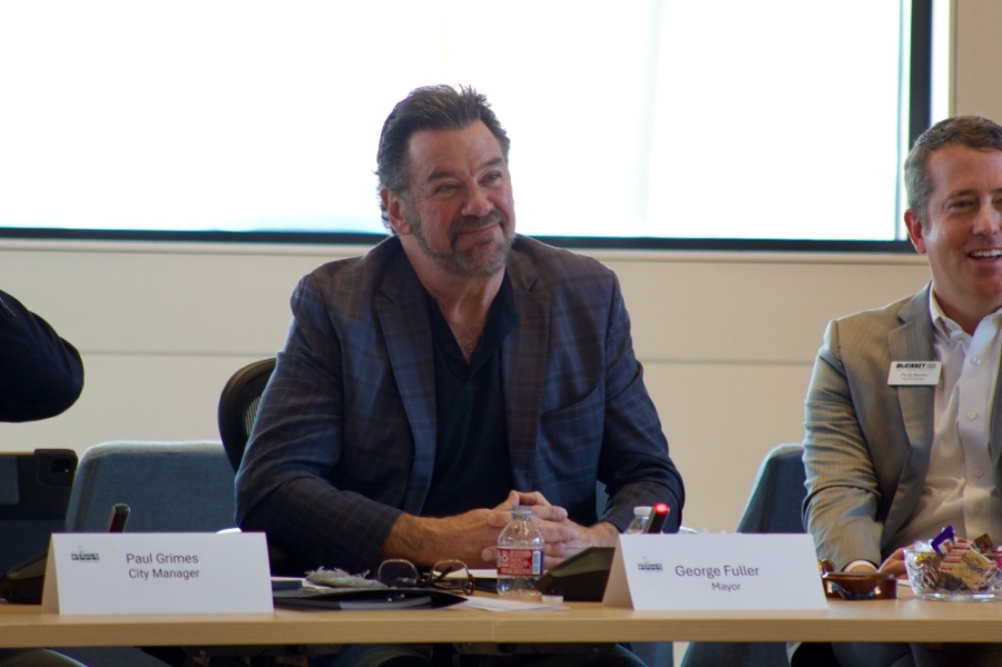 Person seated at a conference table, smiling, between name plates labeled Paul Grimes, City Manager, and George Fuller, Mayor.