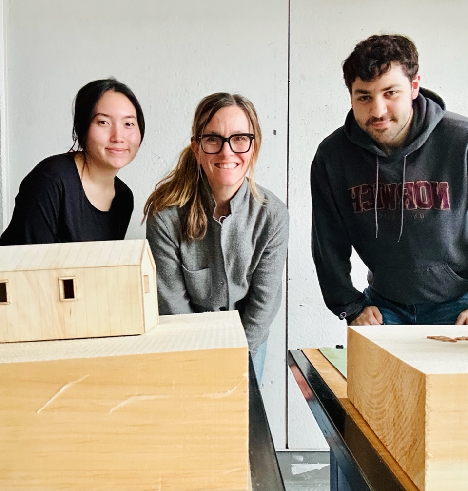 Three individuals standing behind architectural models, smiling at the camera in a workshop setting. The person on the right is wearing a Norwich University sweatshirt.
