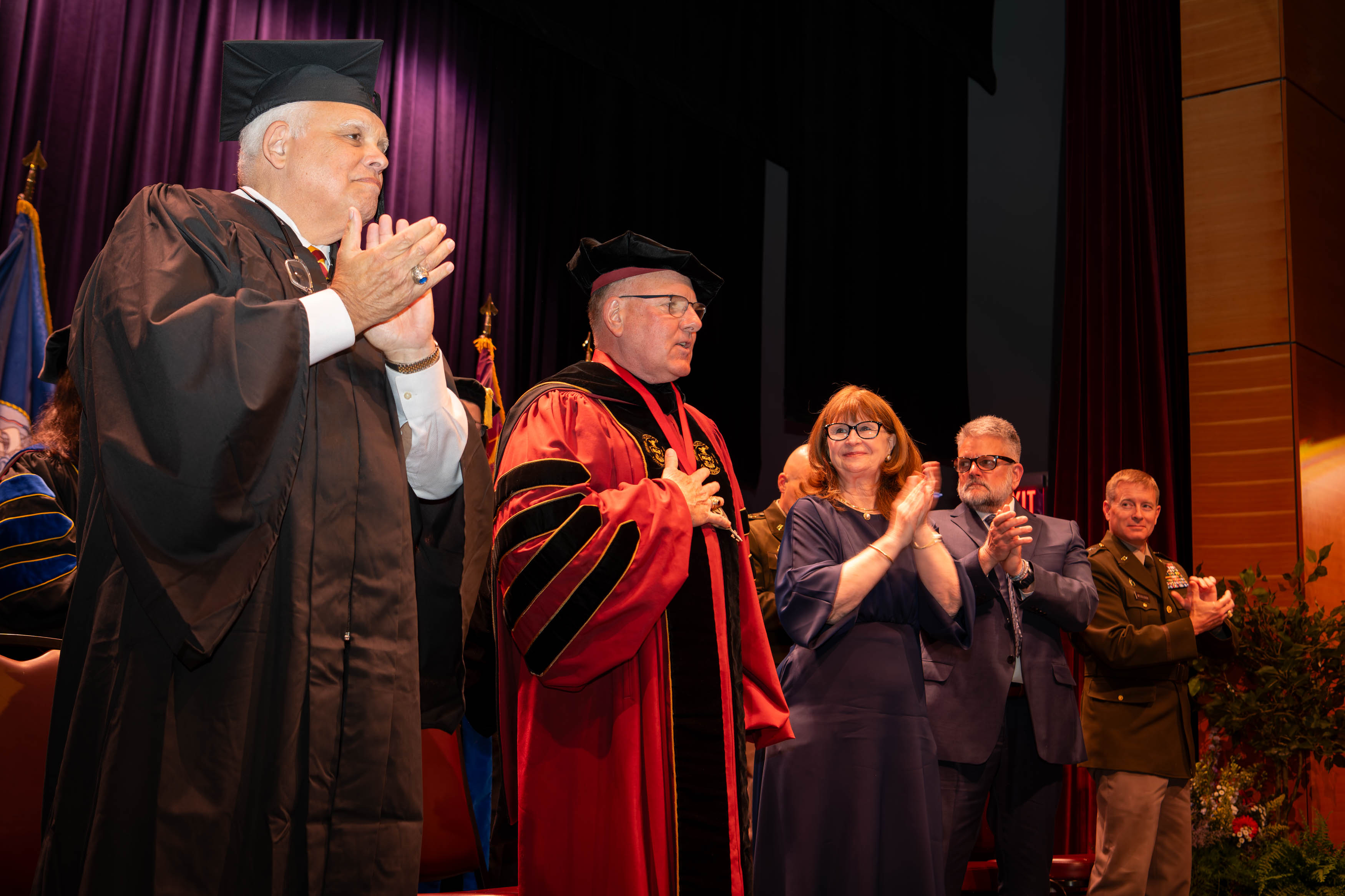 John J. Broadmeadow, University president, stands in the center of the stage in full academic regalia with hand over heart. Chairman Alan DeForest is on his right, while his wife Karen is on his left.