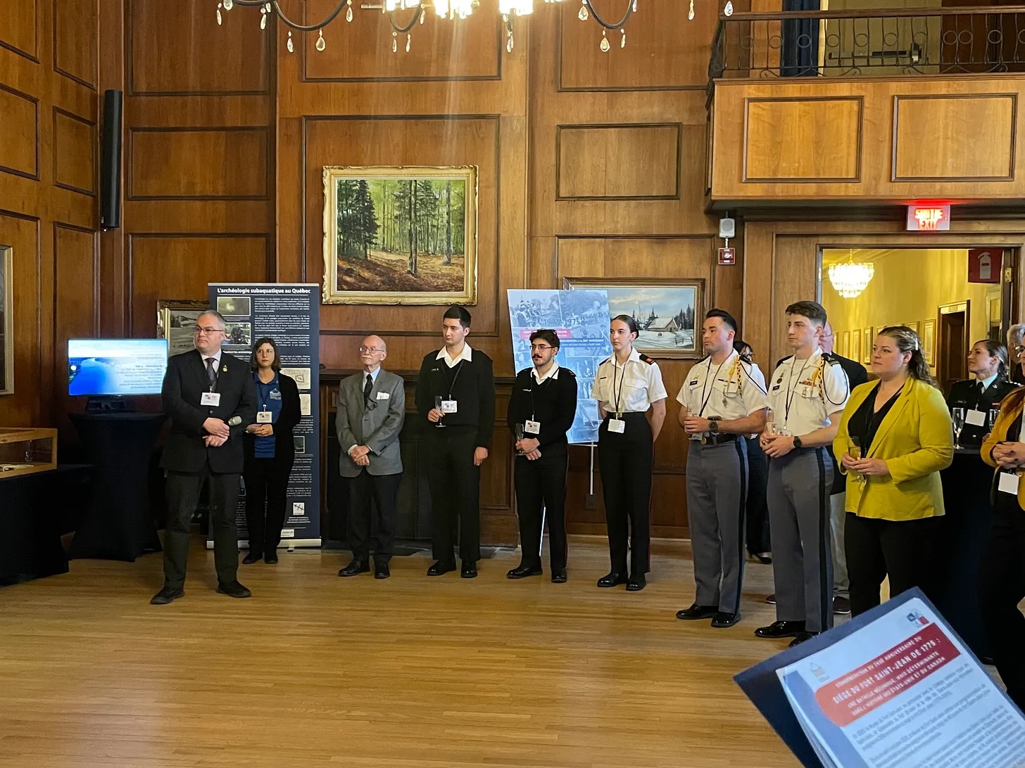 Group of individuals in professional attire standing in a wood-paneled room, with paintings and informational posters on display.