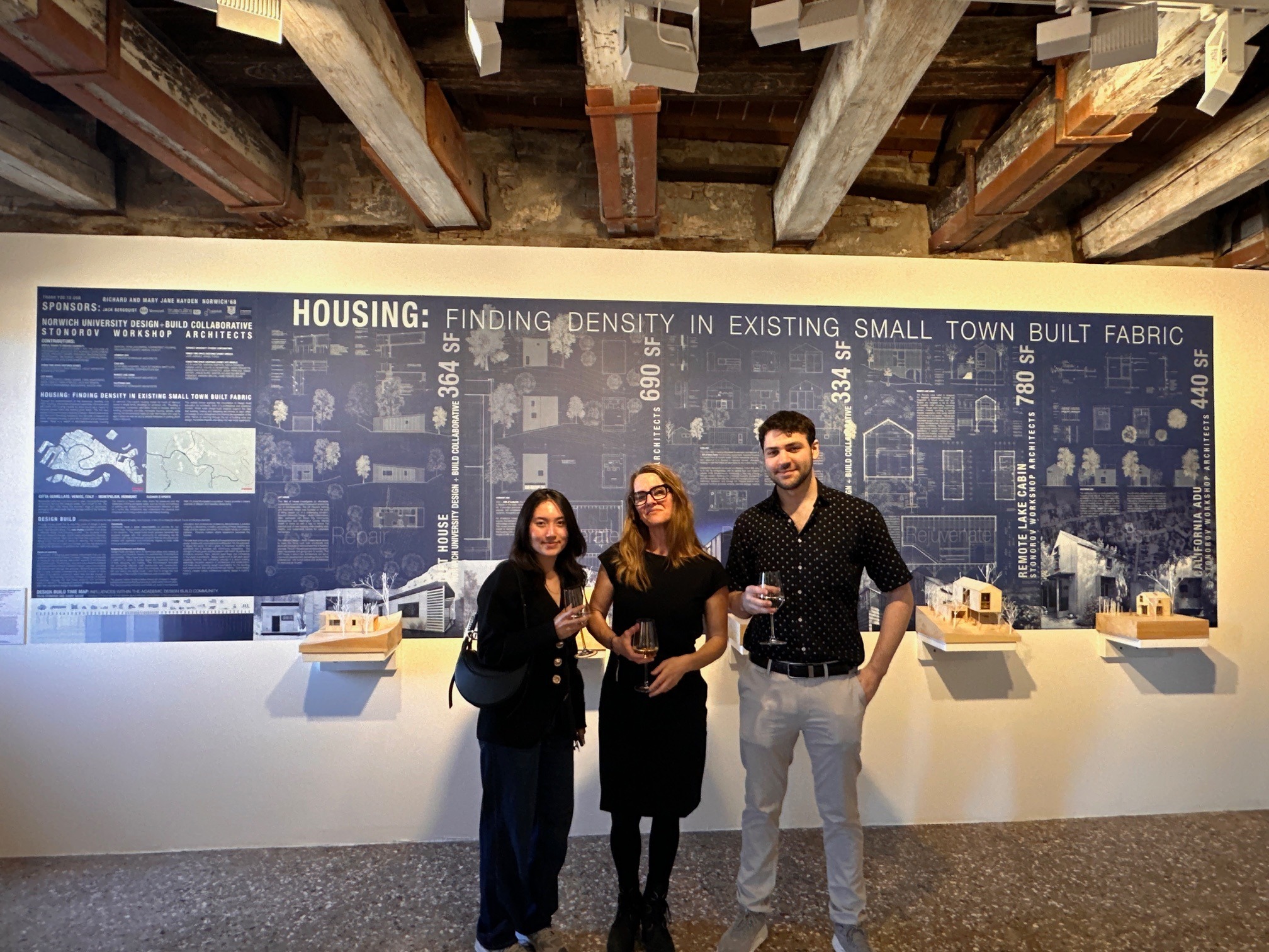 Three individuals holding drinks stand in front of an exhibition panel titled "HOUSING: FINDING DENSITY IN EXISTING SMALL TOWN BUILT FABRIC" at an architectural gallery. They are smiling.