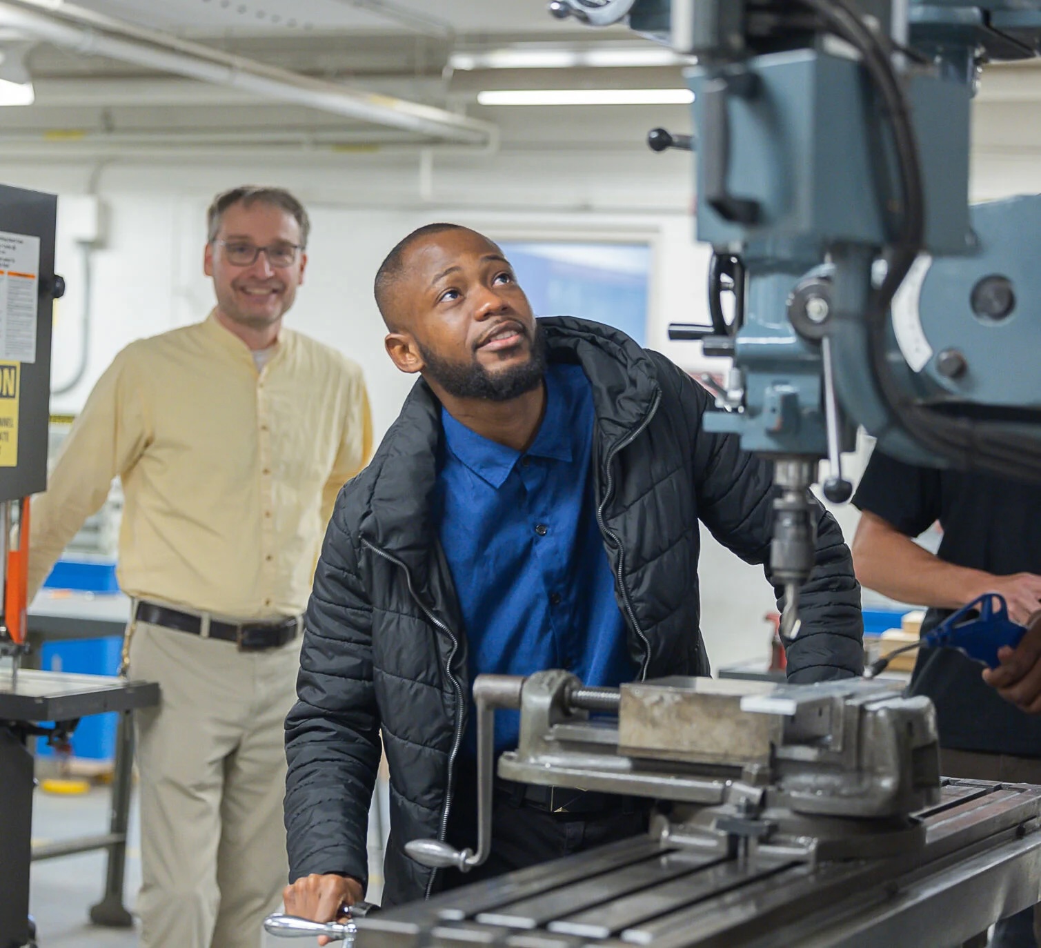 An individual operating a vertical milling machine in an industrial setting while another person observes in the background.