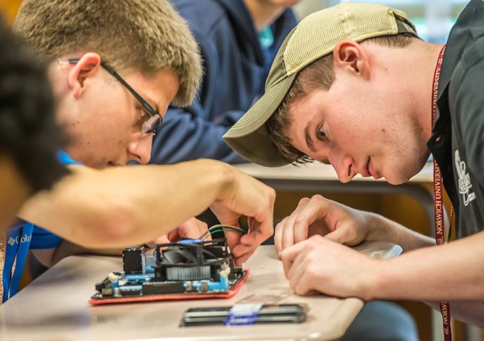 Two Norwich University students work with focused concentration, assembling electronic components on a circuit board at a classroom table.