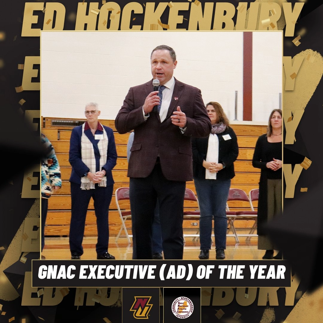 Person speaking at a microphone during an event in a gymnasium, with the banner reading "Ed Hockenbury GNAC Executive (AD) of the Year" visible in the foreground. Several other individuals stand in the background.