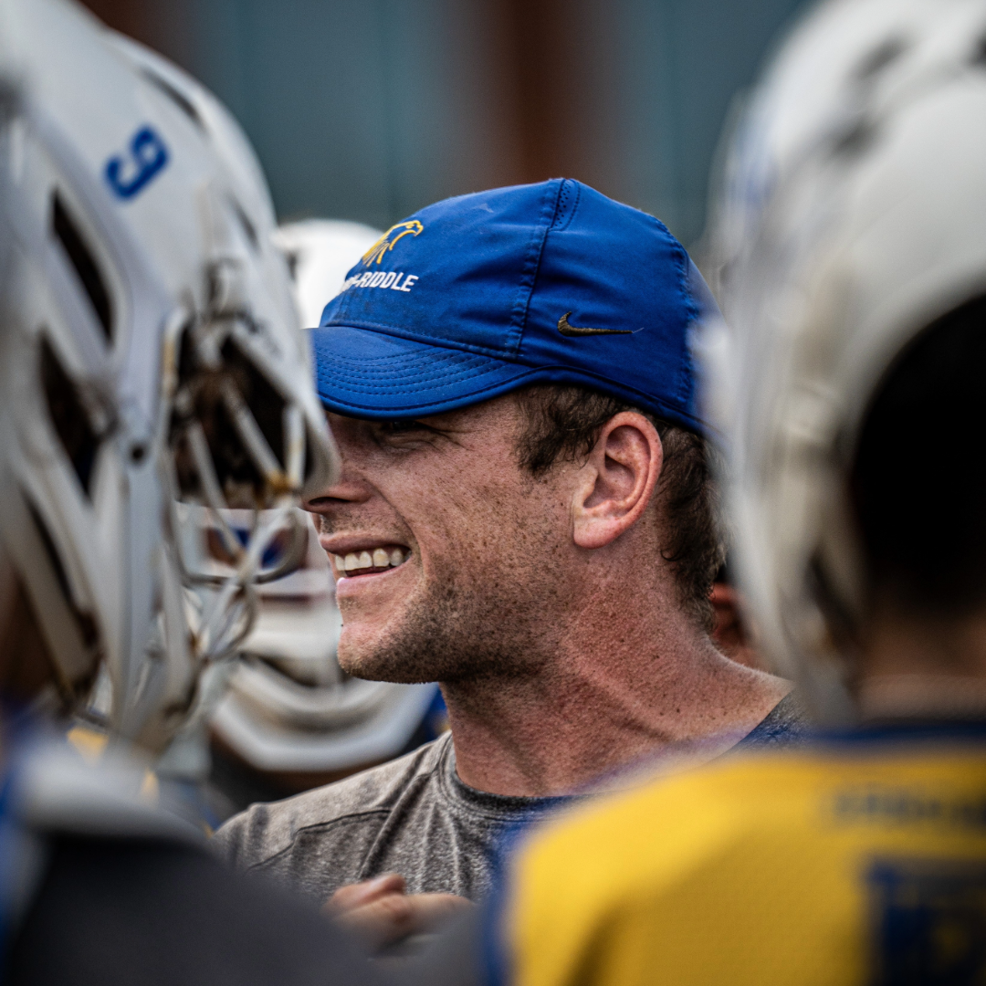 A coach wearing a blue cap smiles while surrounded by players in lacrosse helmets.