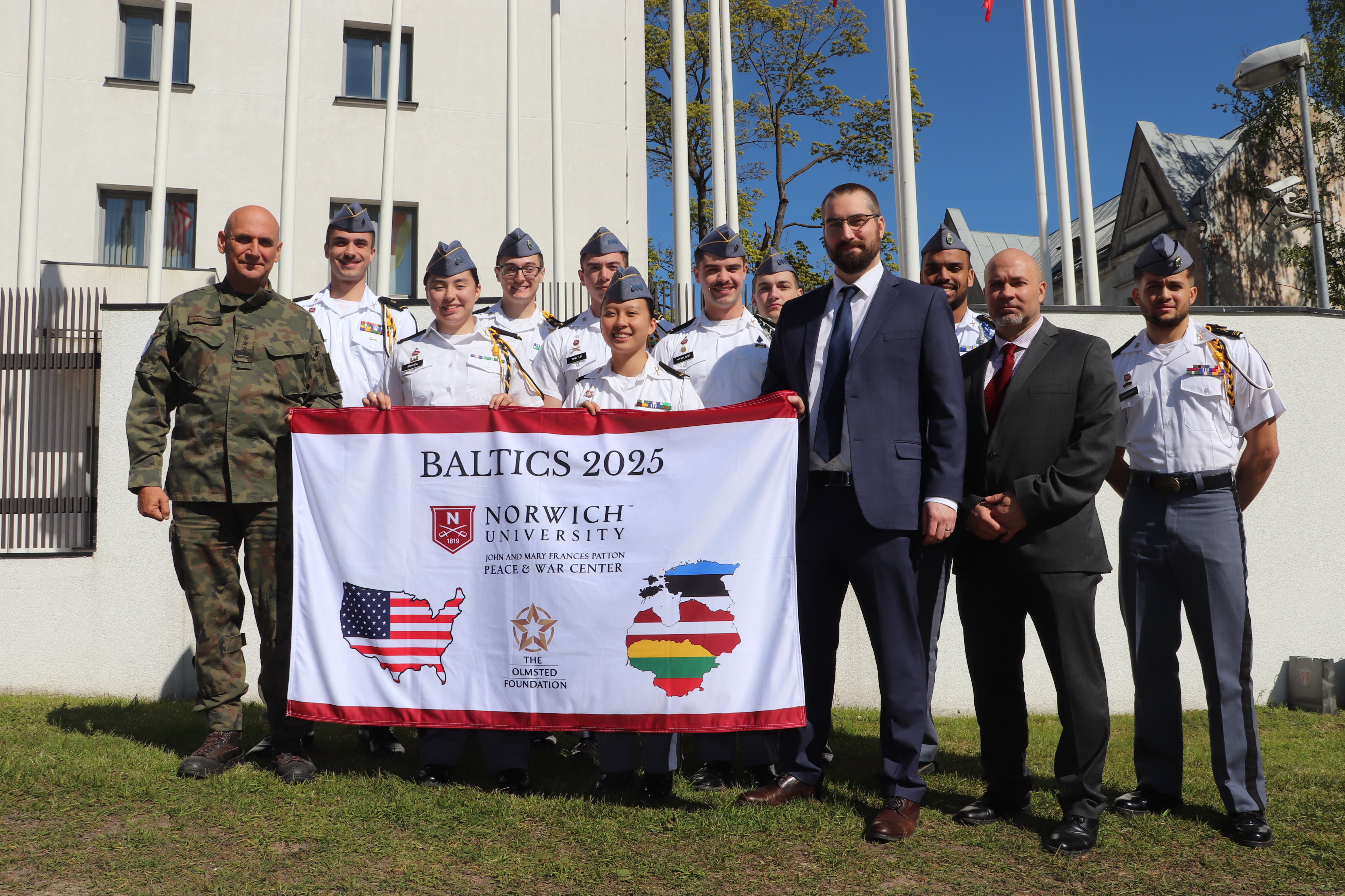 A group photo of the Olmsted trip group, holding a branded Norwich/Olmsted flag in uniform in front of military academy.