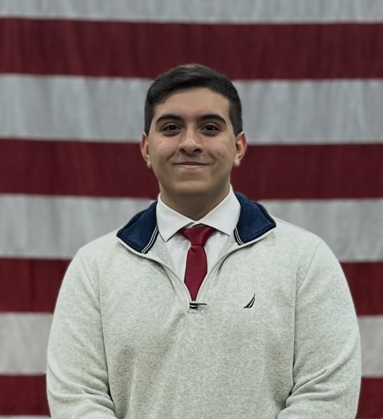 A portrait headshot of Hector Aponte in a sweater, shirt and tie in front of American flag.
