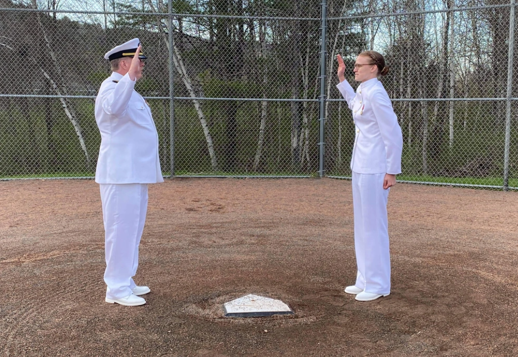Caitlin Dailey (right) stands next to home plate while her father (left, directly across home plate) swears her in. They are both in full white Navy outfits.