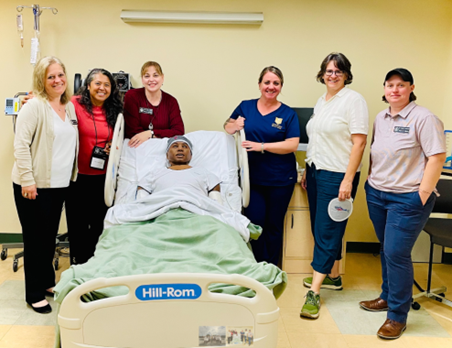 (Left to Right) Jennifer Coutu, Rea Colby, Janie Szalajeski-Hull, Amy Martone, Olga Ryan, and Cindy Freudenthal pose with a high-fidelity 3G manikin, Thomas, in one of the acute care hospital rooms located in the School of Nursing Clinical Simulation Center.