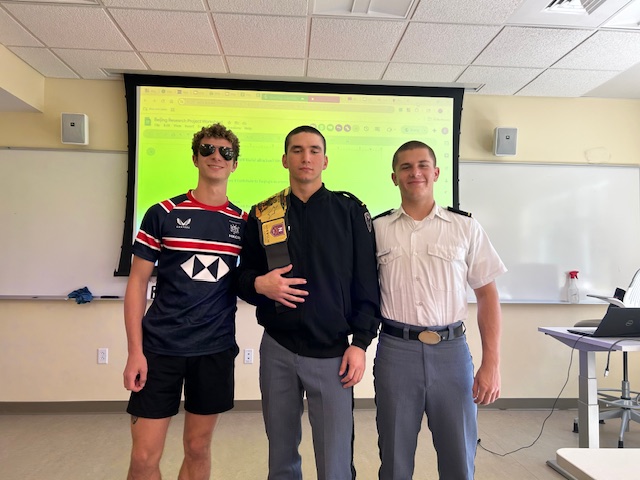Three young men are standing together in a classroom, posing for a photo. The person on the left is wearing sunglasses and a sports jersey, the person in the middle is in a dark jacket with a patch on the shoulder, and the person on the right is wearing a white uniform shirt with gray trousers, typical of a military or cadet setting. A projection screen and whiteboards are visible in the background, suggesting this was taken during or after a class or presentation.