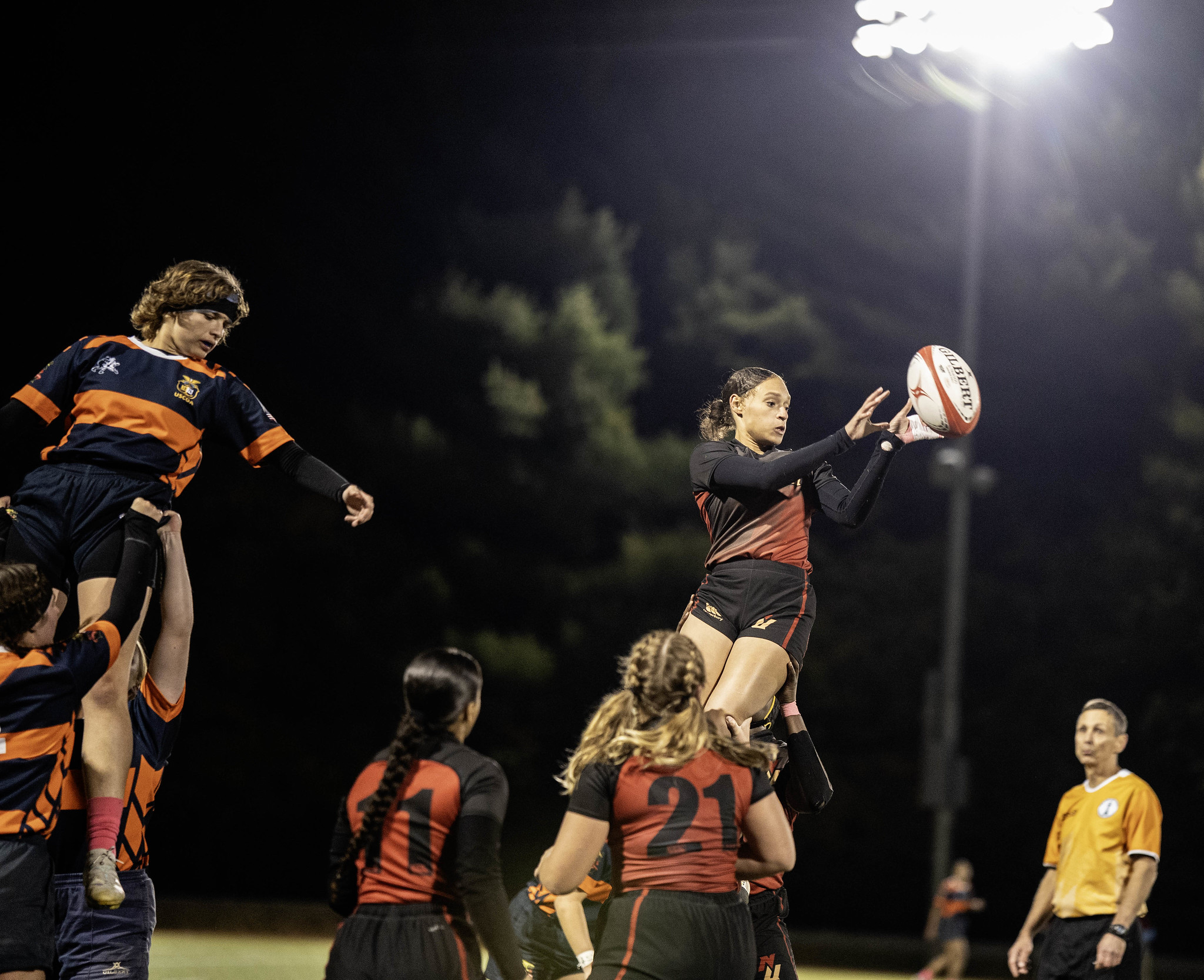 A women's rugby player is lifted into the air while holding the ball during a night match.