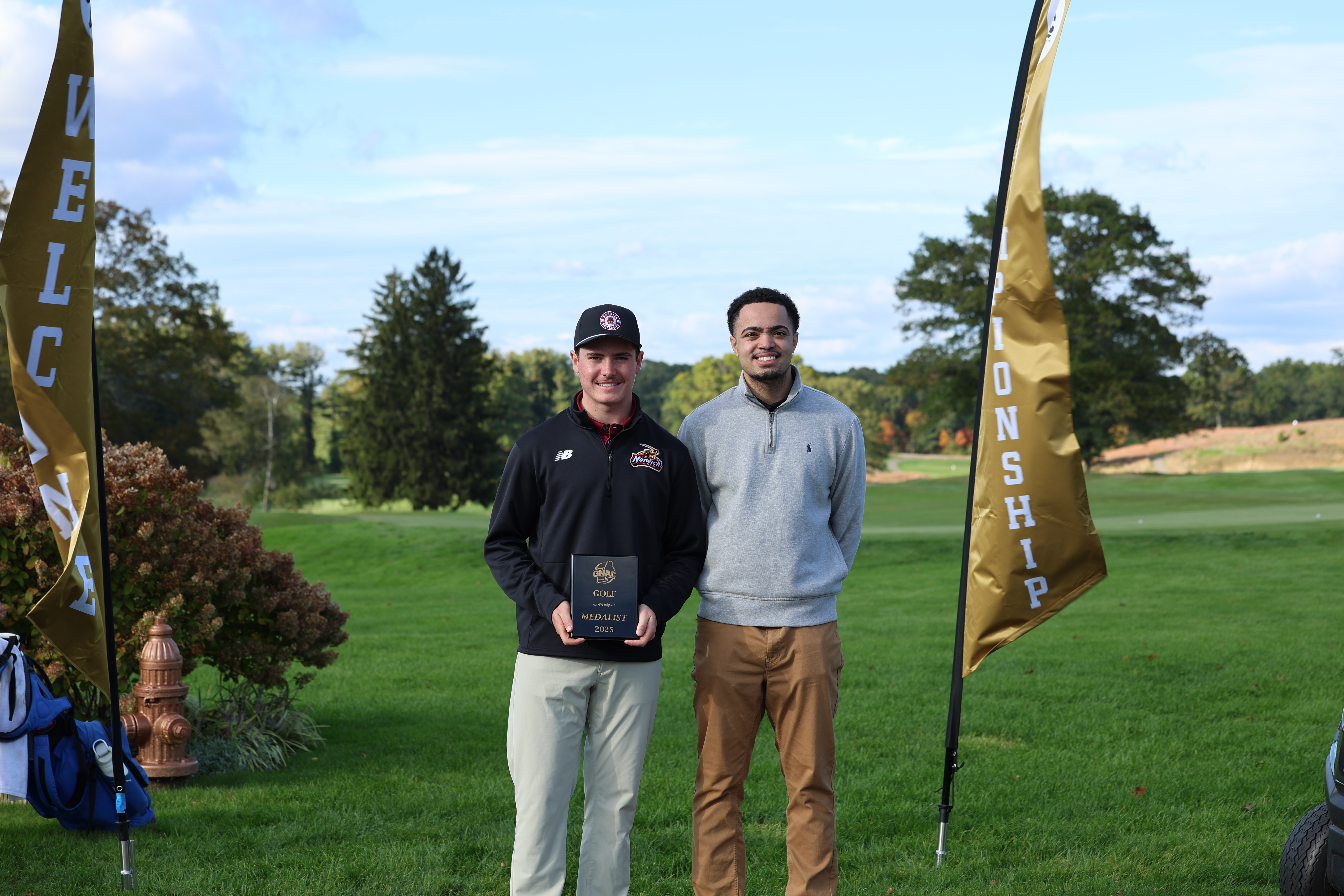 Two people stand on a golf course, one holding a plaque between two gold flags.
