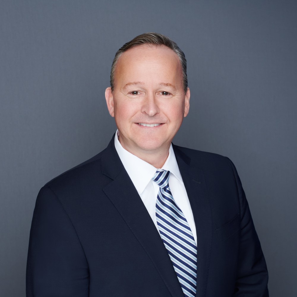 Person wearing a dark suit and striped tie poses against a gray background.