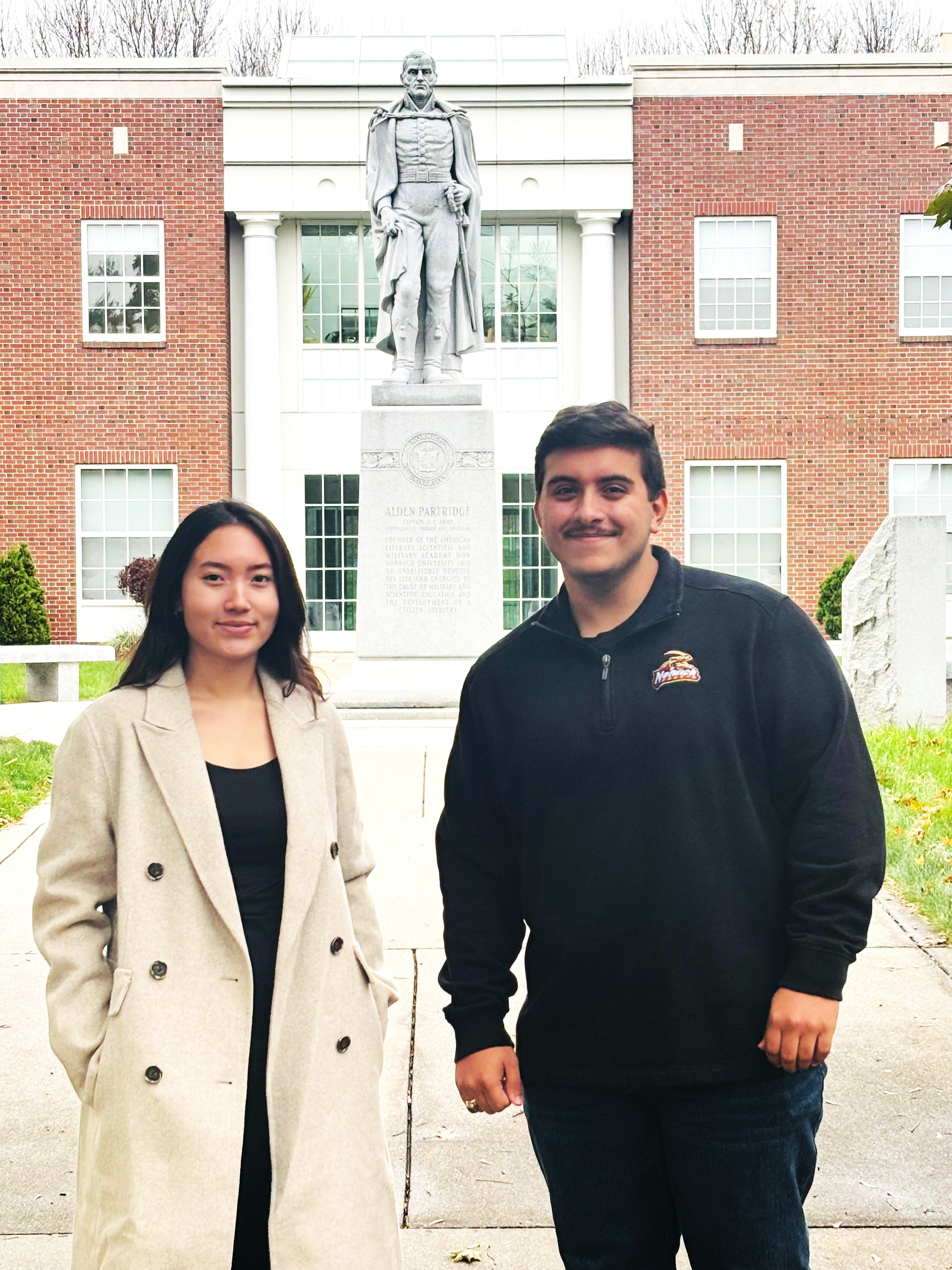 Two people stand outside near a statue in front of a red brick building with white columns.