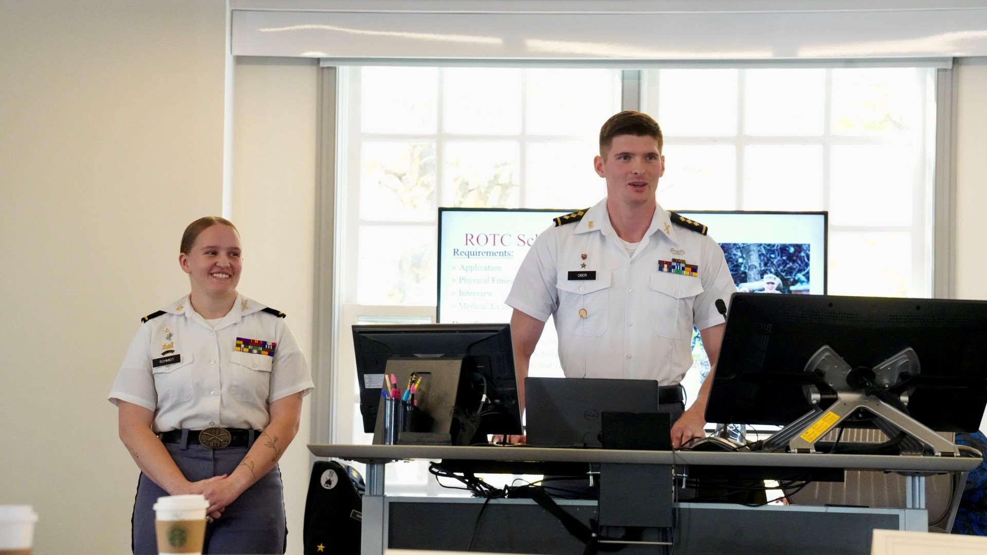 Two cadets in uniform are standing at the front of a classroom giving a presentation. One is speaking while the other smiles and listens. Both are wearing white shirts with rank insignia and ribbons. Behind them, a screen displays a slide with “ROTC Scholarship Requirements” listed. The environment appears professional and academic, with computers, notes, and coffee cups visible on the desks.