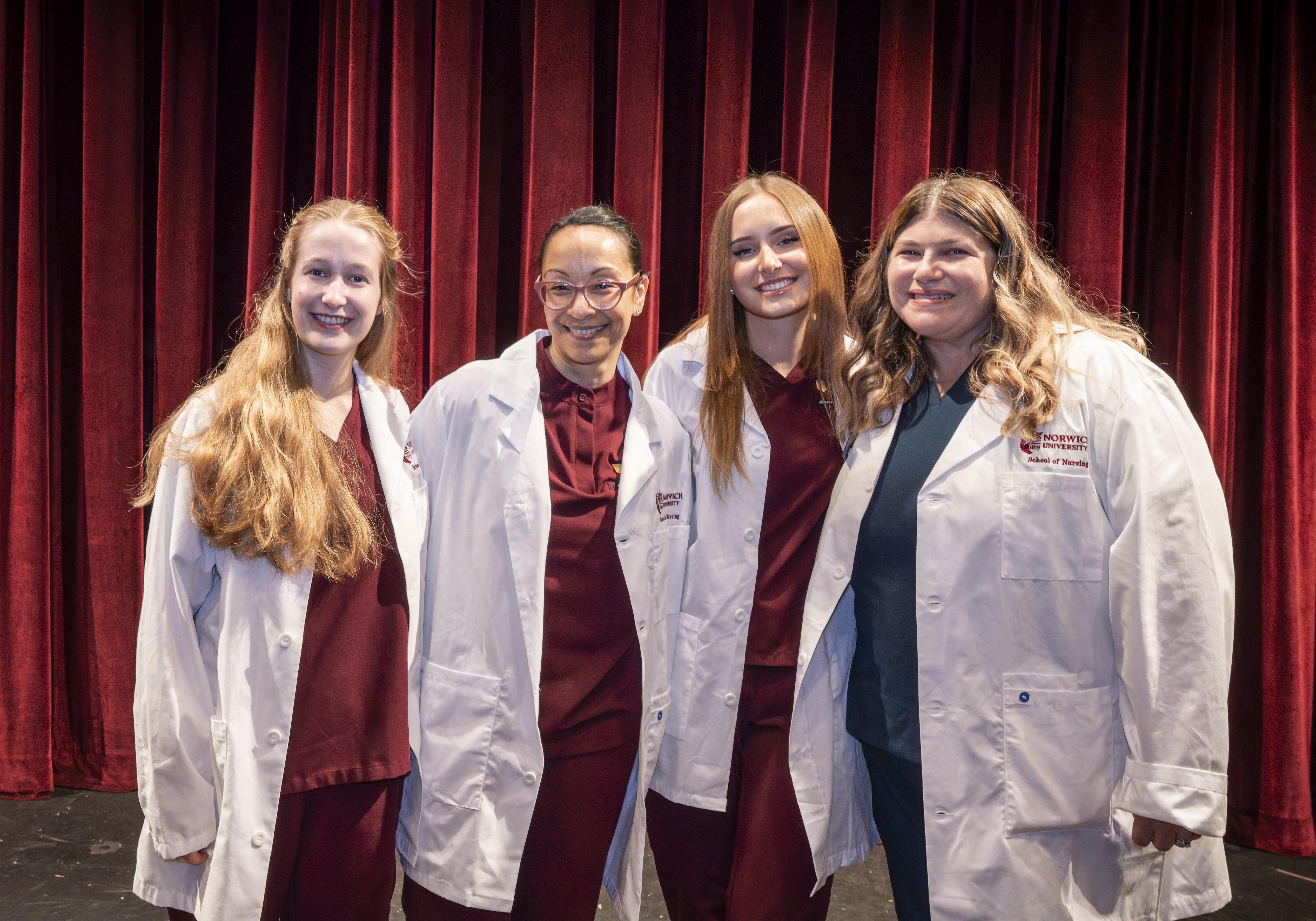 Four individuals stand together smiling, dressed in maroon scrubs and white coats embroidered with “Norwich University School of Nursing.” The backdrop of rich red curtains and stage lighting gives the setting a formal and celebratory tone.