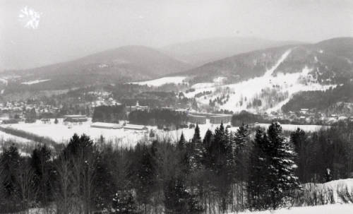 A snowy valley stretches across the scene, surrounded by forested hills and distant mountains. A small cluster of buildings sits nestled near the center, with open fields and ski trails visible along the slopes.