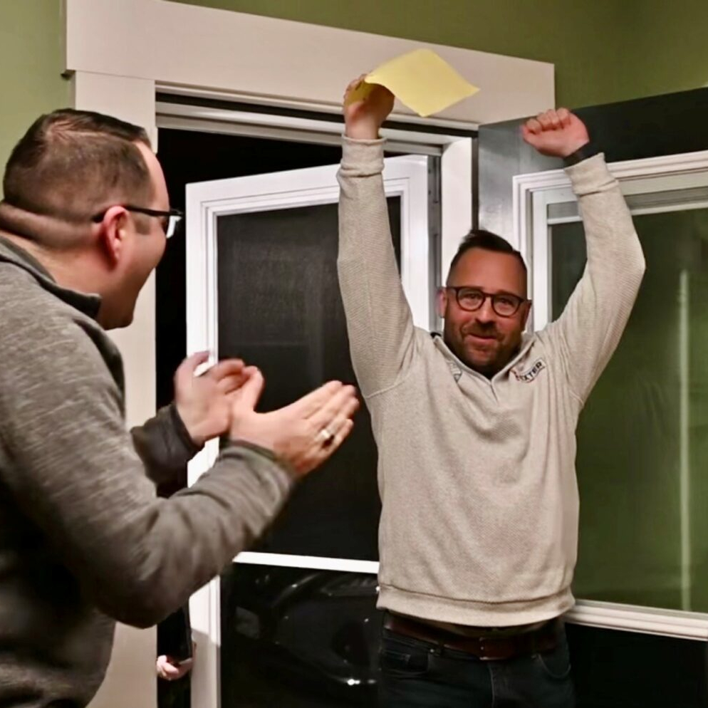 a man in foreground, facing another man with hands in air, holding a paper ballot