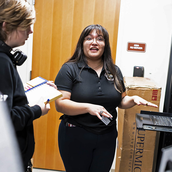 A woman stands in front of a computer server, instructing an observer