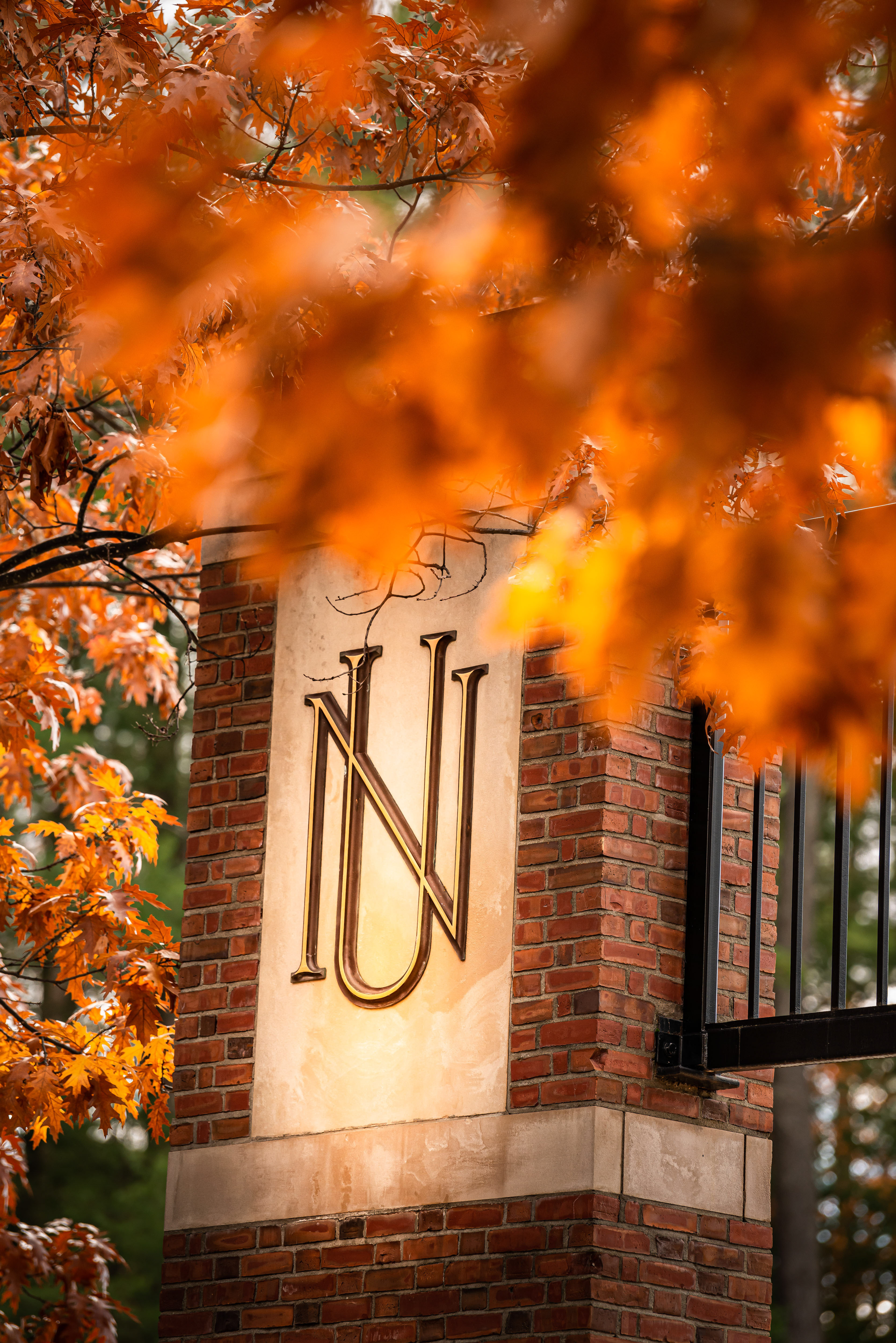 A brick pillar with the logo "NU" on a gold plaque, framed by vibrant orange autumn leaves.