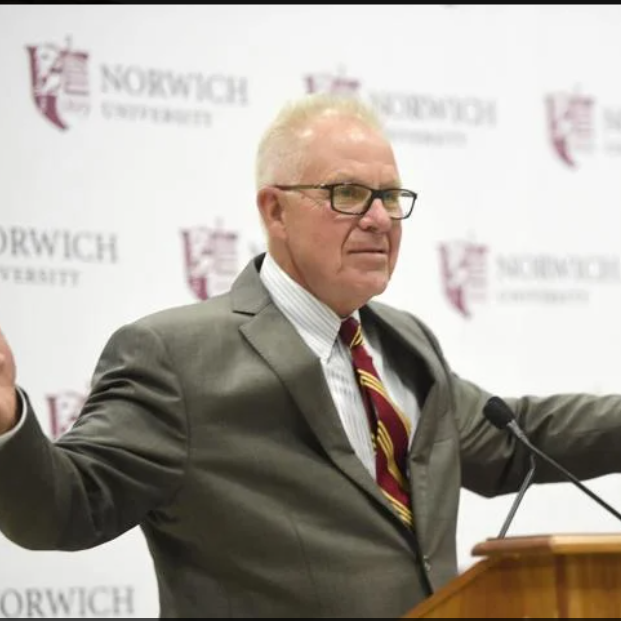 A man stands at a podium with a Norwich University banner behind him.