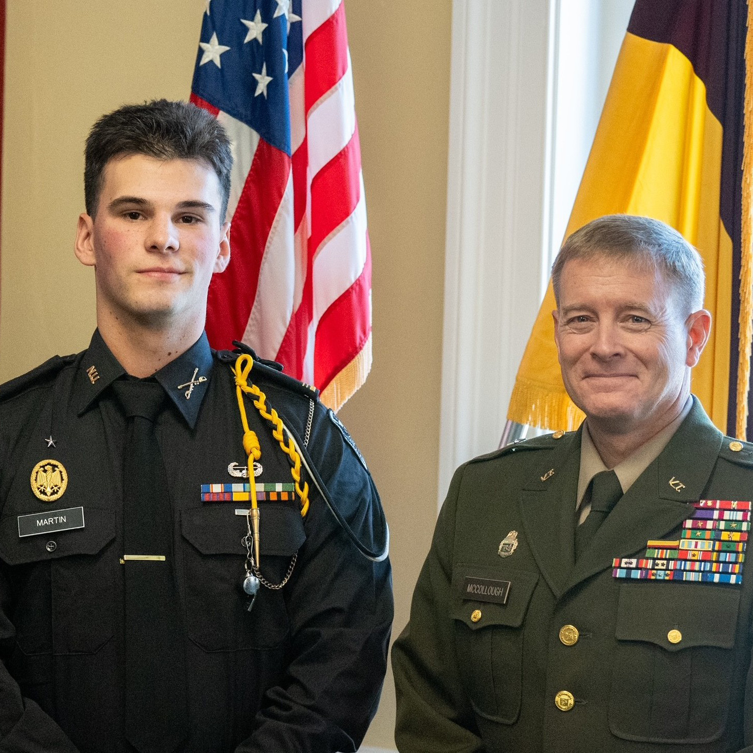 Two people in military dress uniforms stand indoors in front of U.S. and yellow-and-brown flags.
