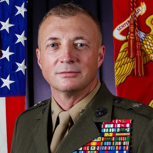 Person in a decorated military uniform stands before U.S. and Marine Corps flags.