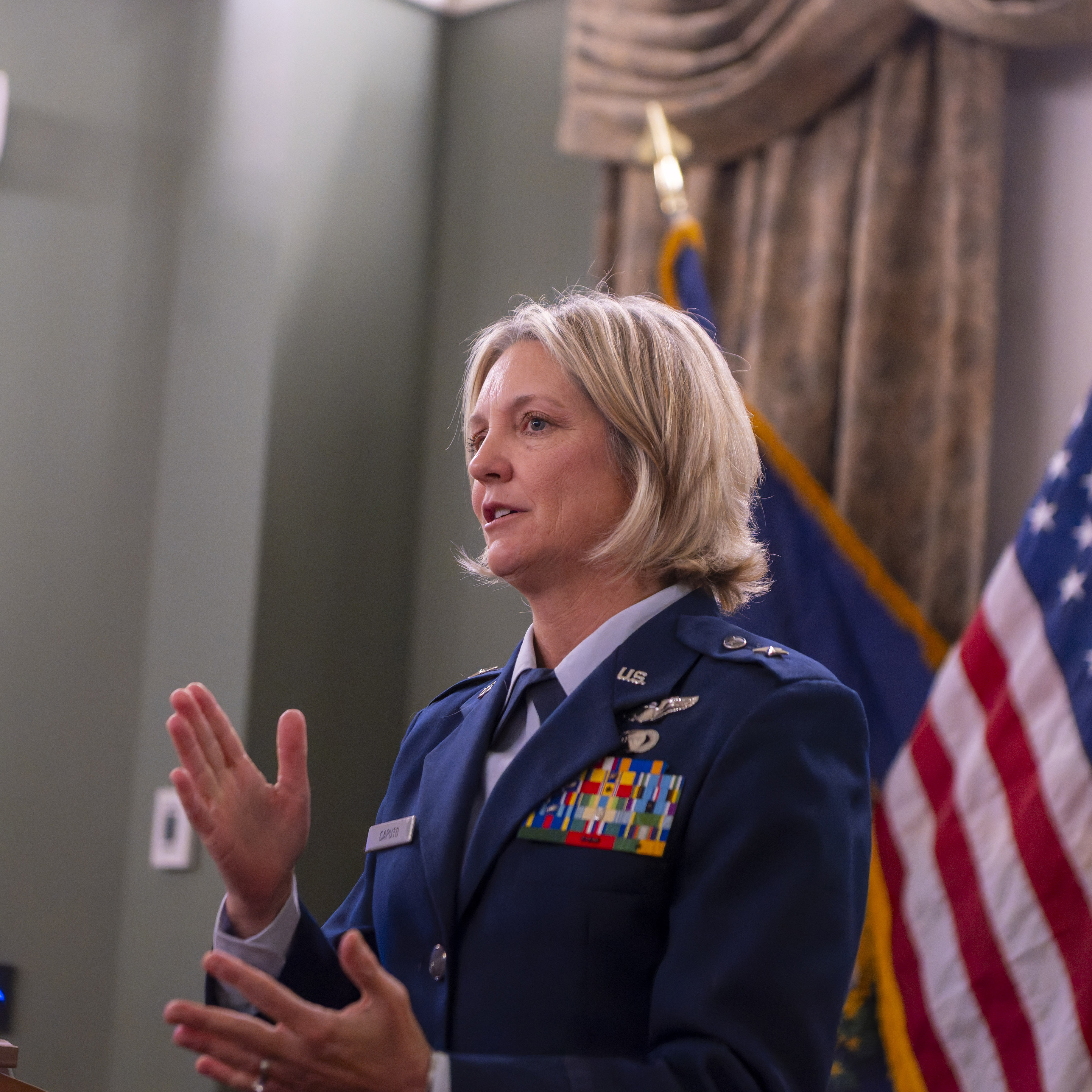Person in a military dress uniform gestures while standing near flags and curtains in a formal room.