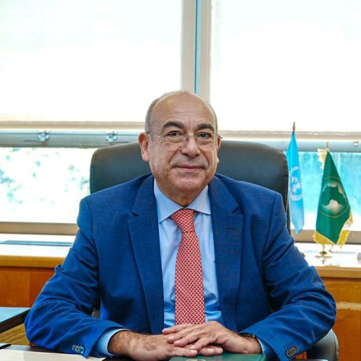 Person in blue suit sits at desk with hands clasped, with two flags and a window behind.