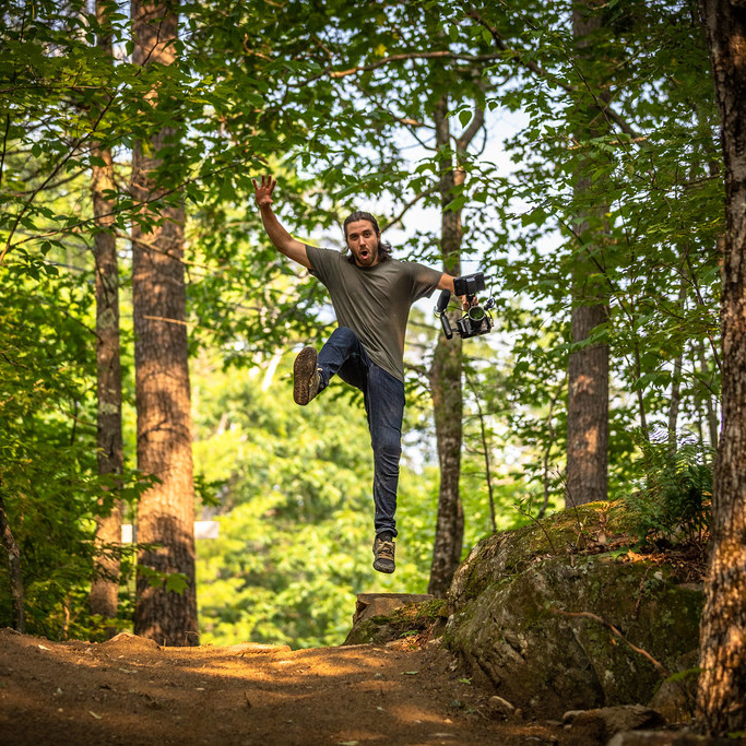 A person jumps mid‑air on a forest trail holding a camera with sunlight filtering through the trees.