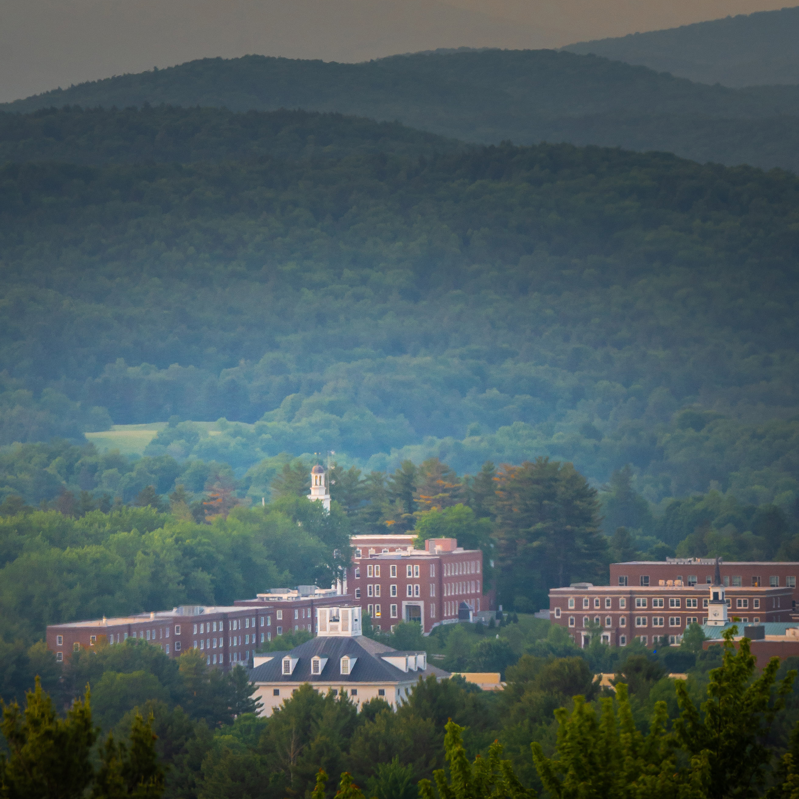 Red brick campus buildings sit among dense green trees with layered forested hills in the background.