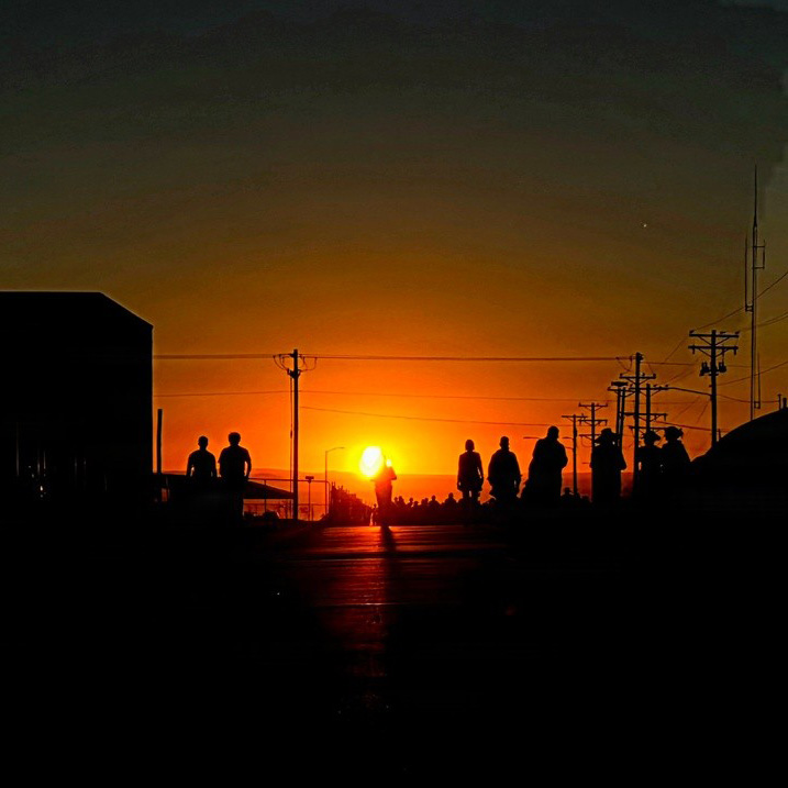 Silhouetted people walk along a road at sunrise with utility poles on both sides.