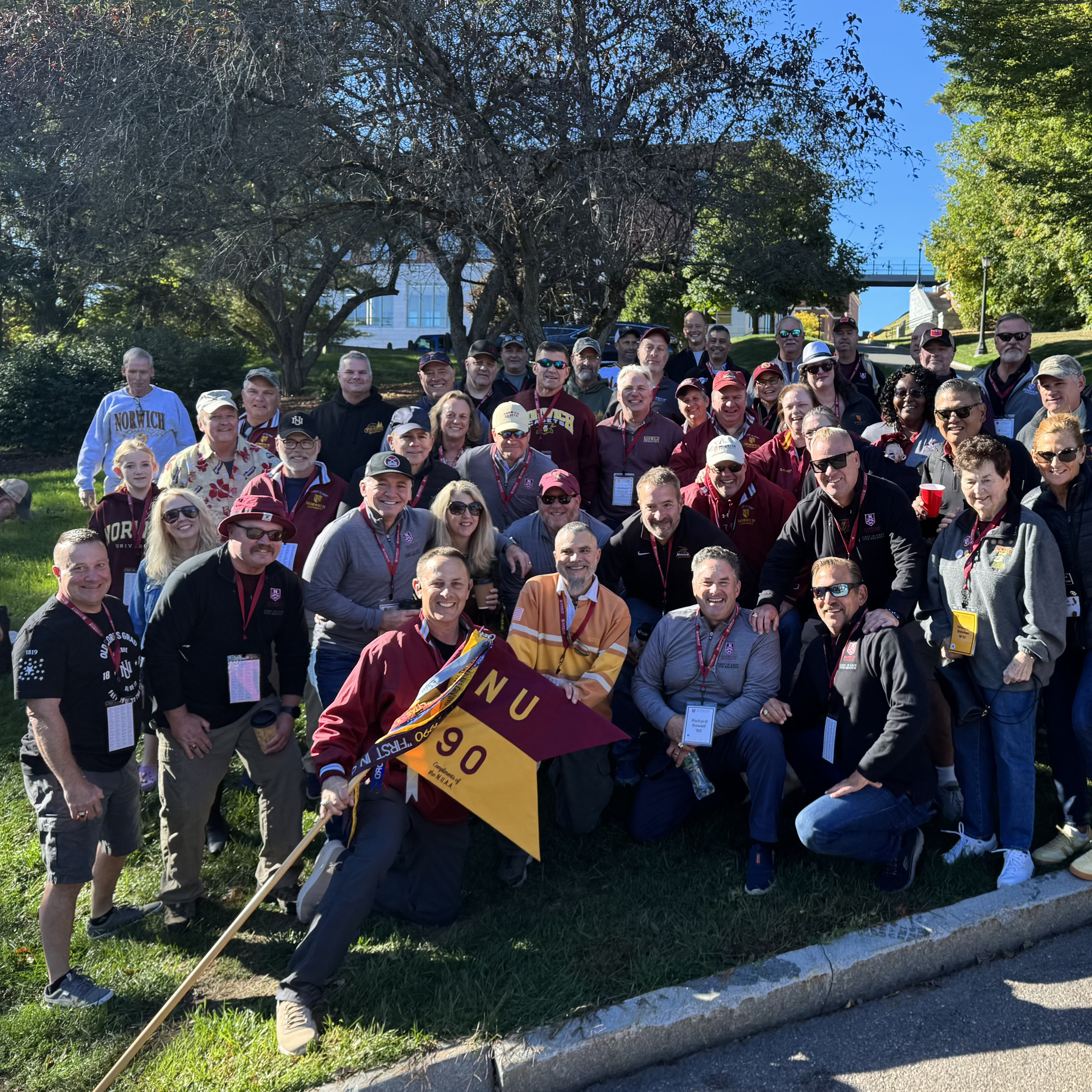 Group of people stand on a lawn holding a Norwich NU 90 flag near trees and a campus walkway.