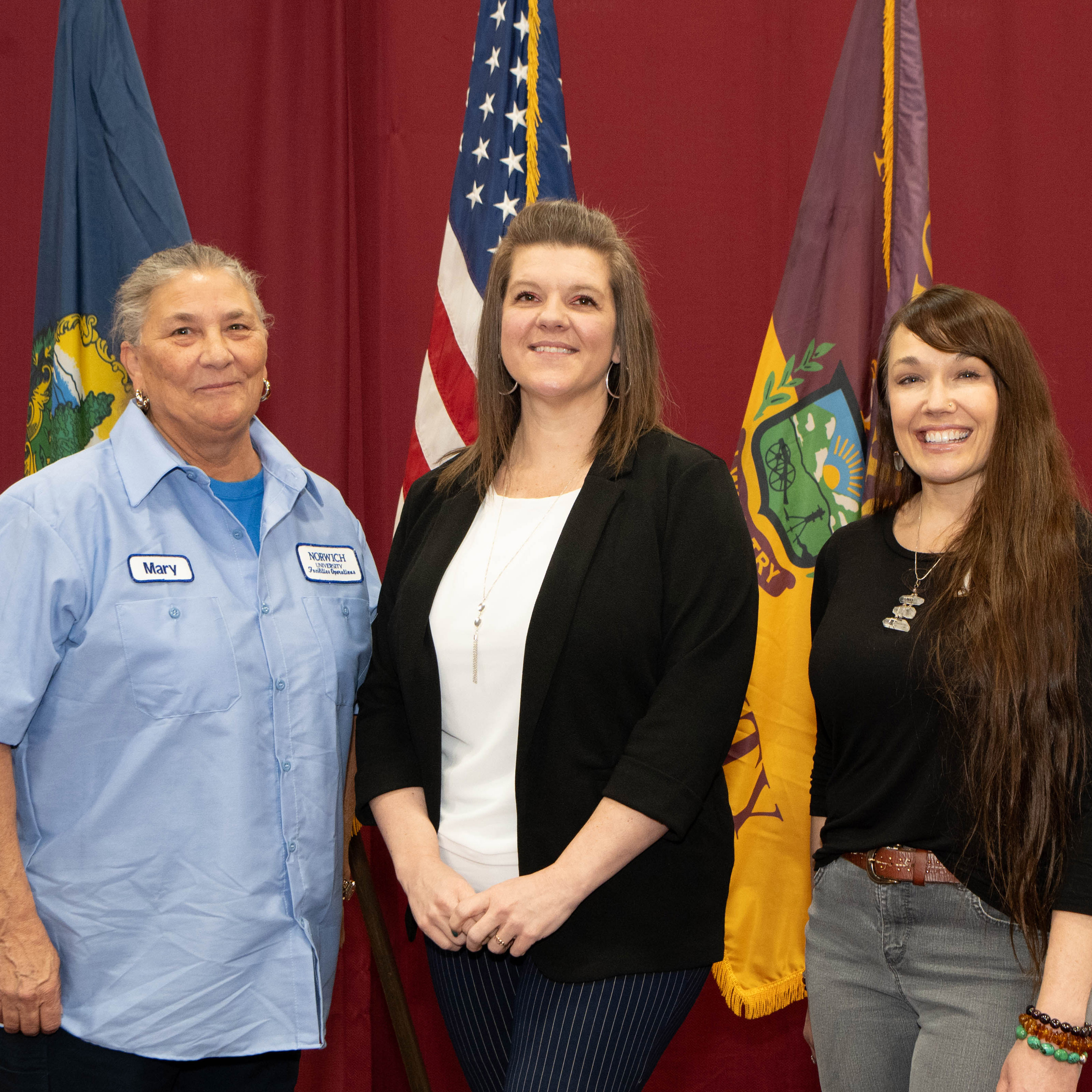 Three staff award recipients hold Norwich University certificates in front of flags.
