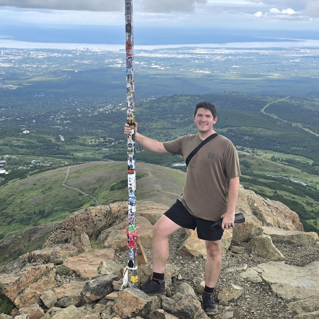 Person stands on rocky summit holding a pole with an American flag above a valley and distant water.