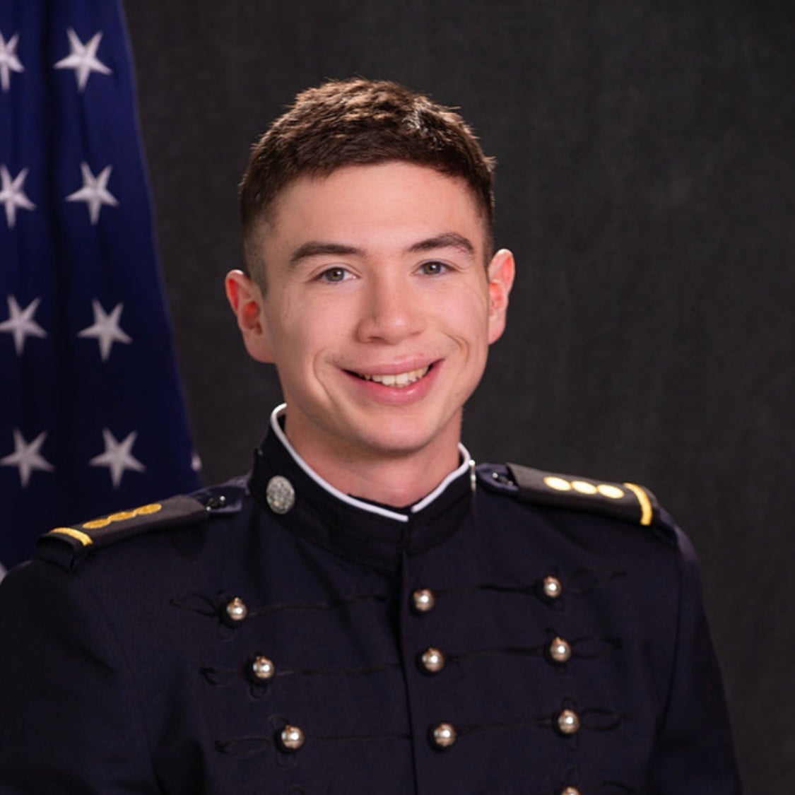 A cadet in blue dress uniform smiles in formal headshot with American flag in background.
