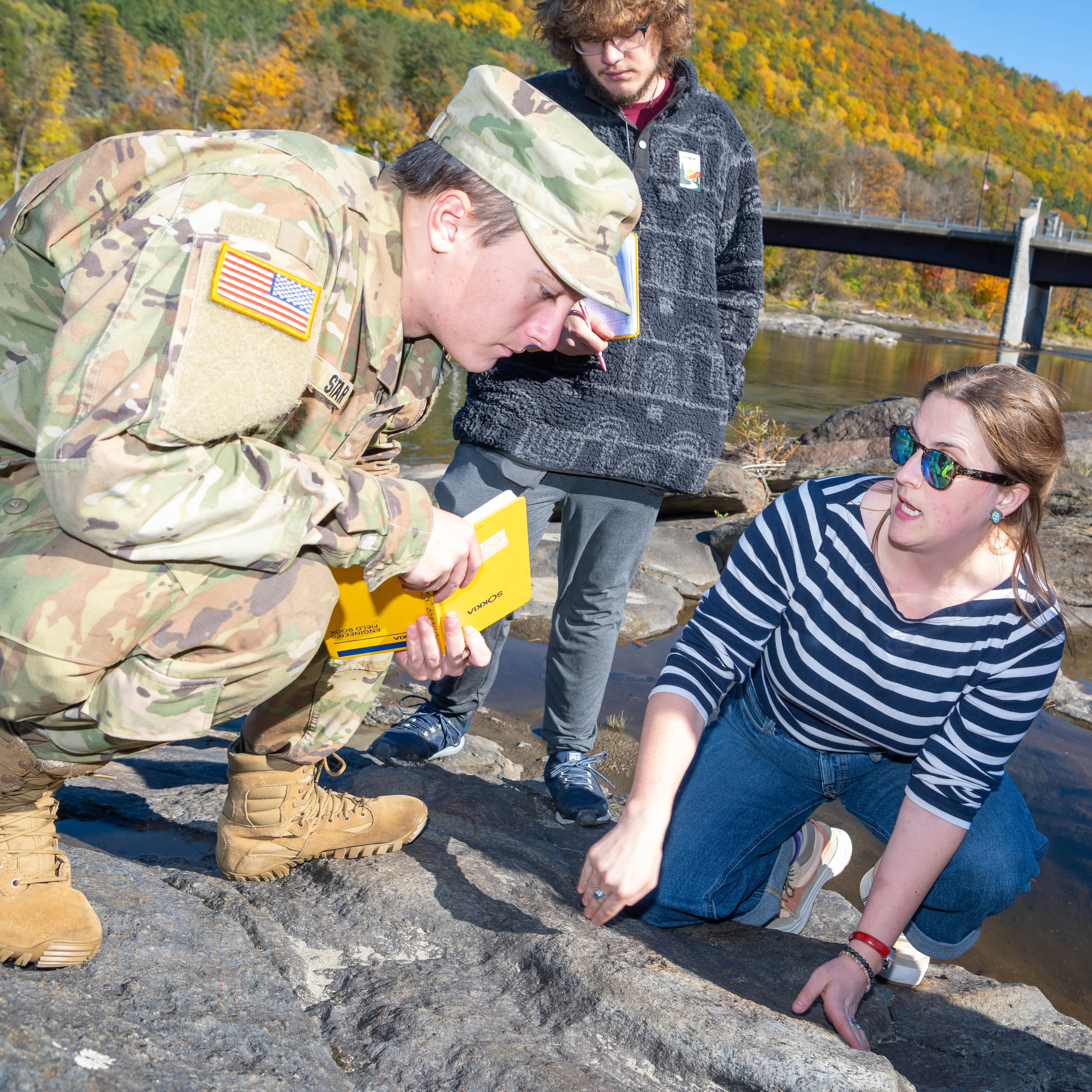 Cadet and students examine rocks beside a river with autumn trees and a bridge in the background.