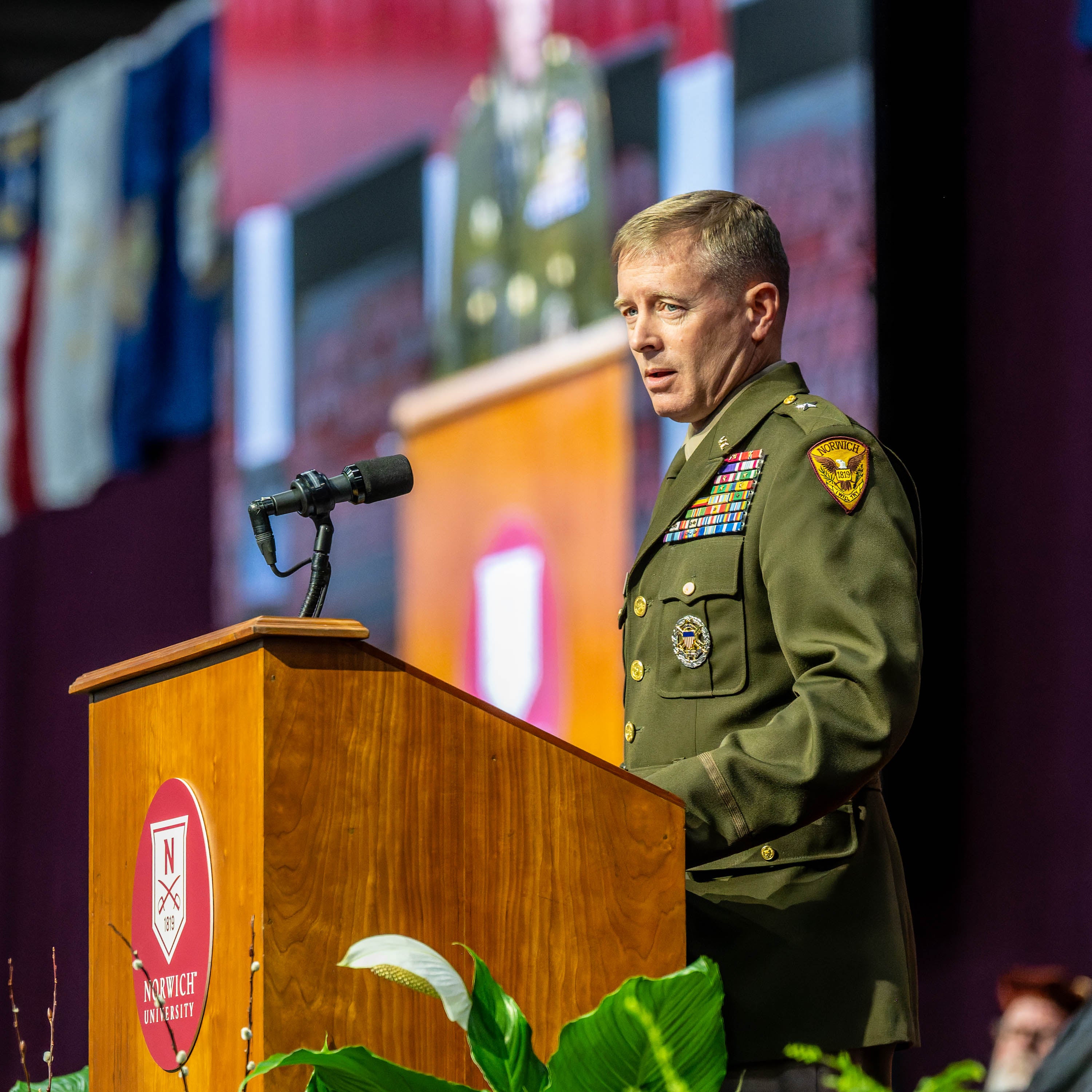 Military officer speaks at Norwich University podium during indoor ceremony.
