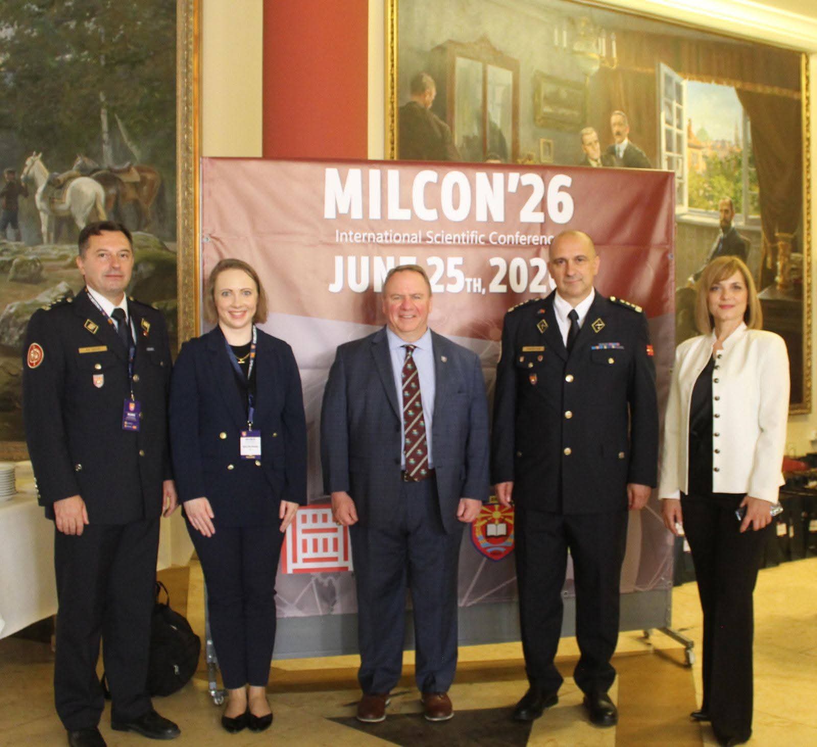 Five people pose in front of a MILCON'26 conference banner inside a room with large framed paintings.