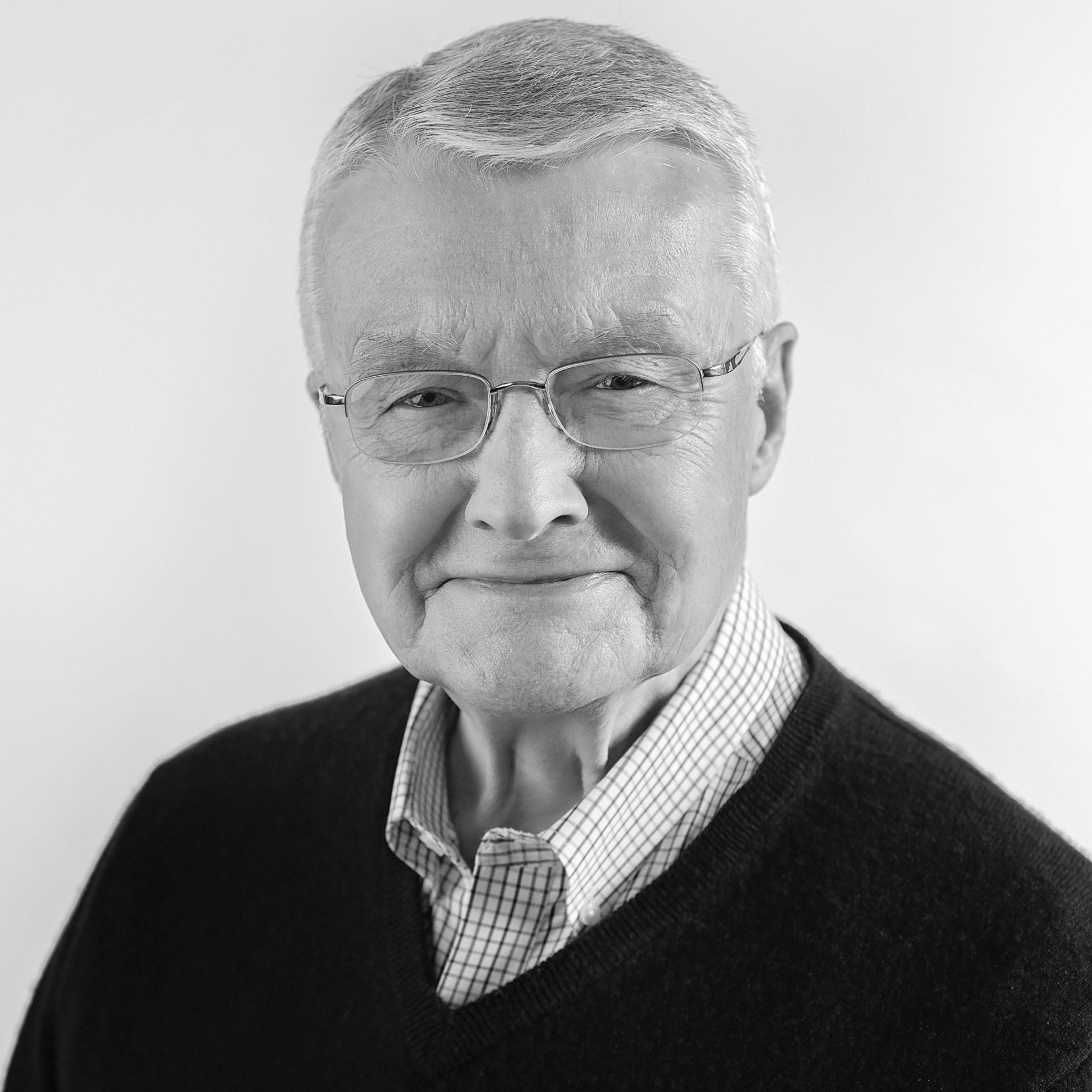 Person with short gray hair and glasses wears a dark sweater over a checkered shirt against a plain background.