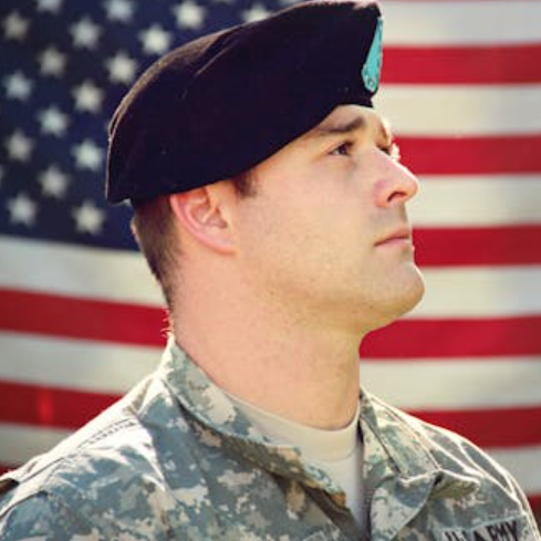 Service member in camouflage uniform and black beret faces right with an American flag in the background.