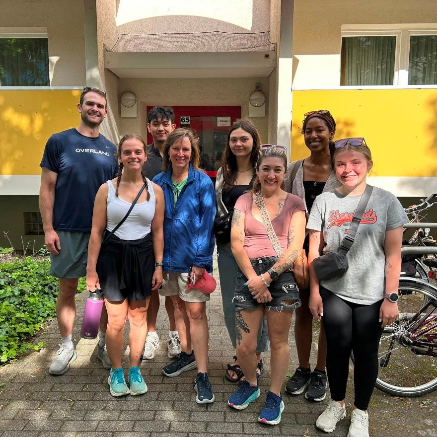 Eight people stand together on a walkway in front of an apartment building entrance with bicycles parked nearby.