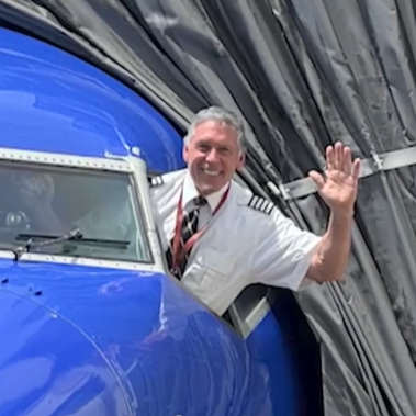 A pilot stands in the cockpit leaning out the open window, waving.