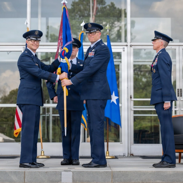 Two service members in blue dress uniforms exchange a ceremonial flag while two others stand nearby outside a building.